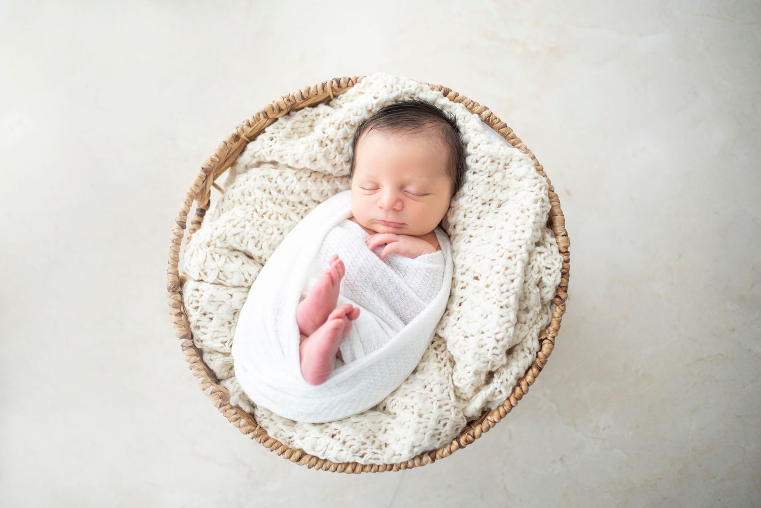 Newborn baby sleeps in round woven basket lined with white textured fabric.