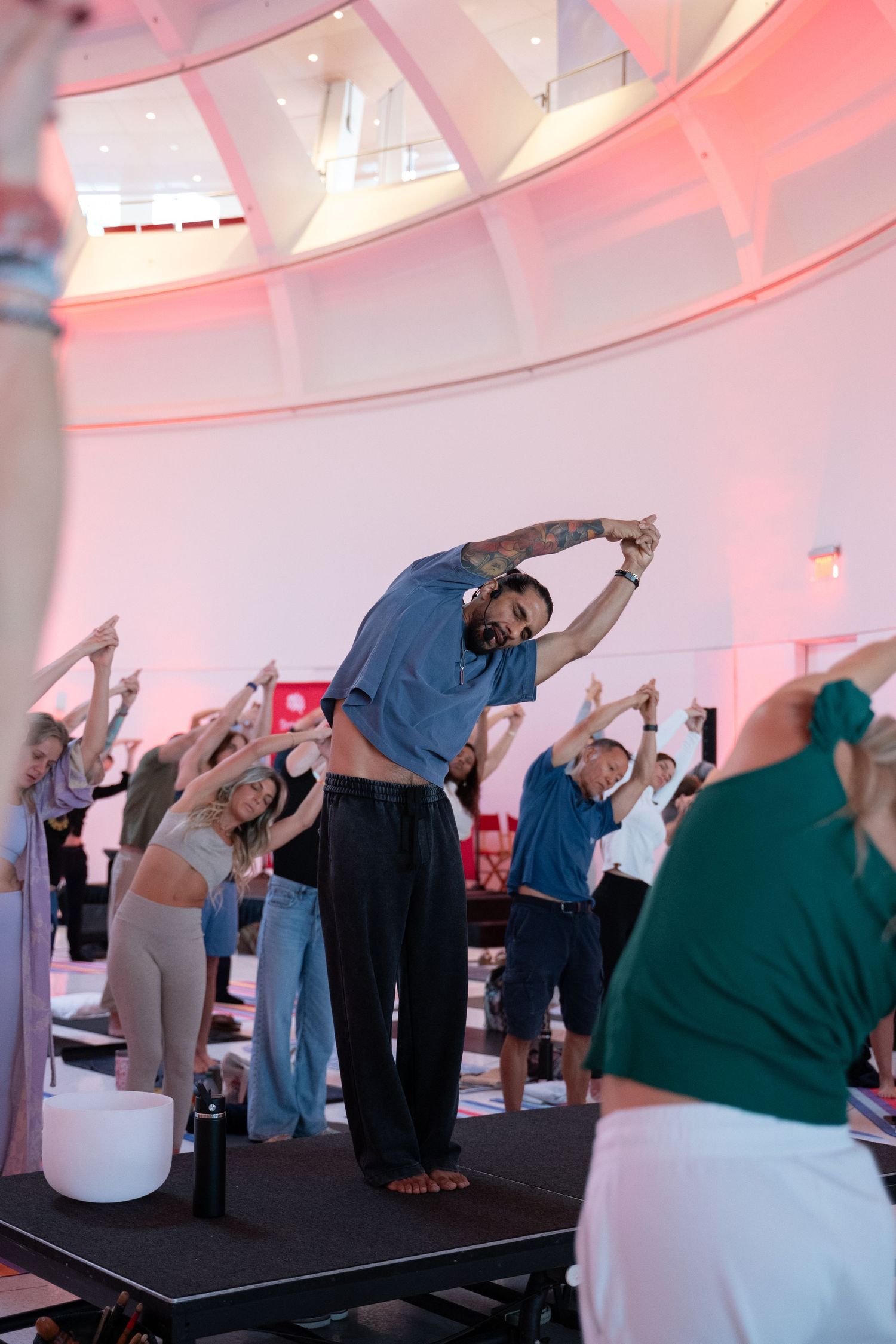 Group yoga class in a modern rotunda venue with pink lighting, participants stretching arms overhead led by tattooed instructor.