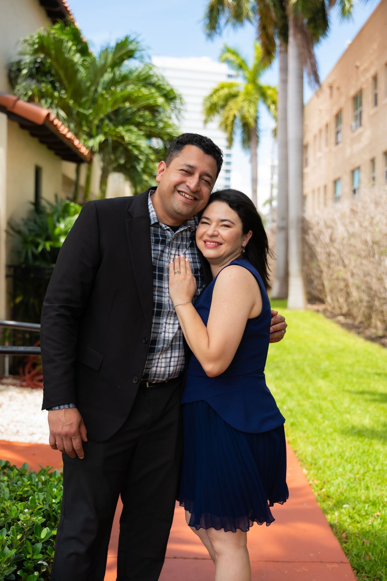 A smiling couple embraces on a red brick path surrounded by palm trees outdoors.