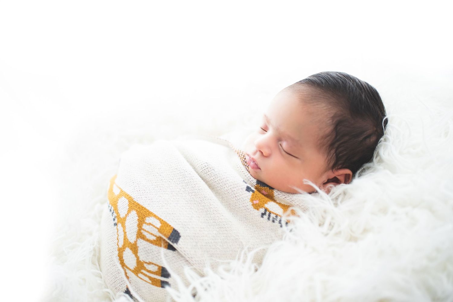 A sleeping newborn baby wrapped in white swaddle blankets lies peacefully on soft white bedding.