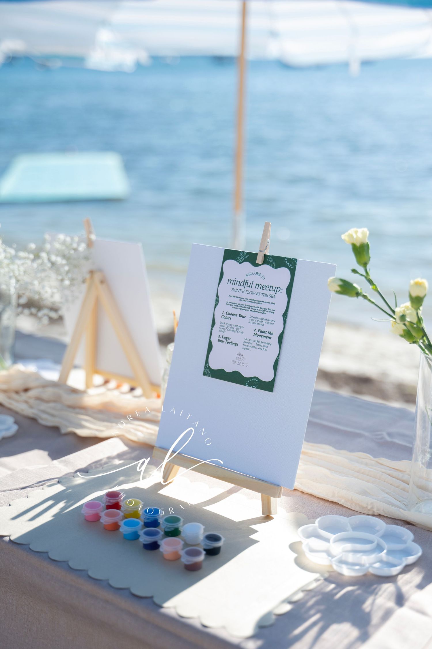 Paint supplies and canvas set up for an outdoor art session on a beach with blue ocean waves in the background.