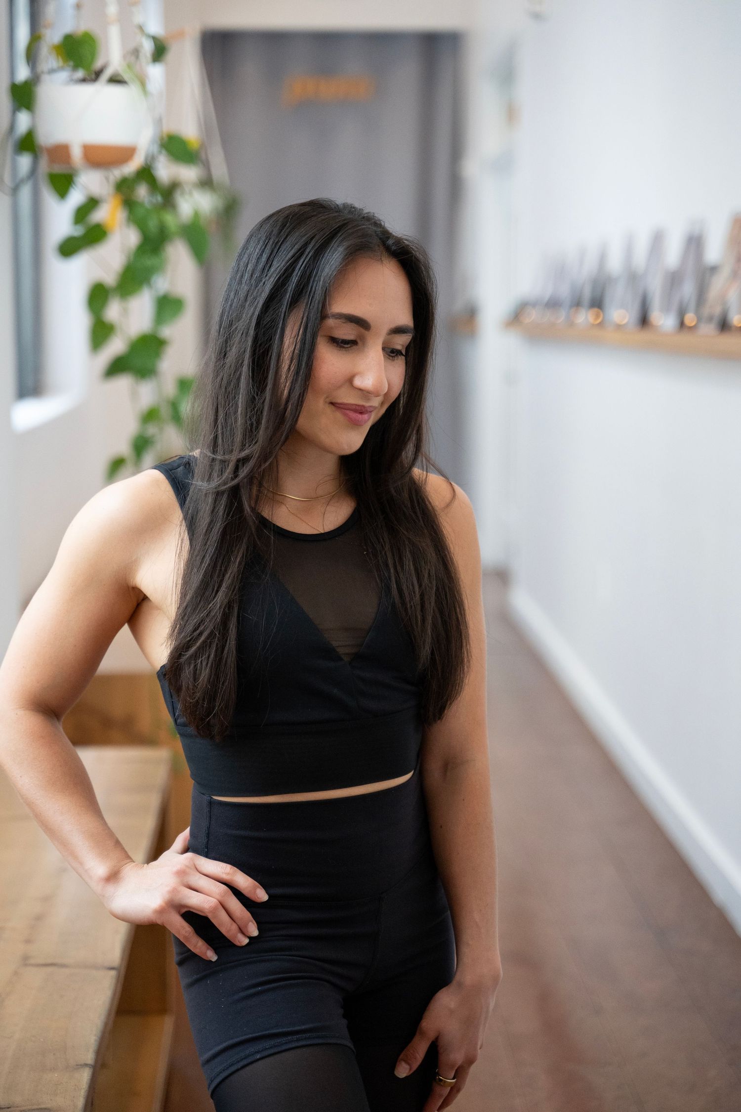 Woman in black athletic wear poses confidently in bright interior space with plants and wooden furniture.