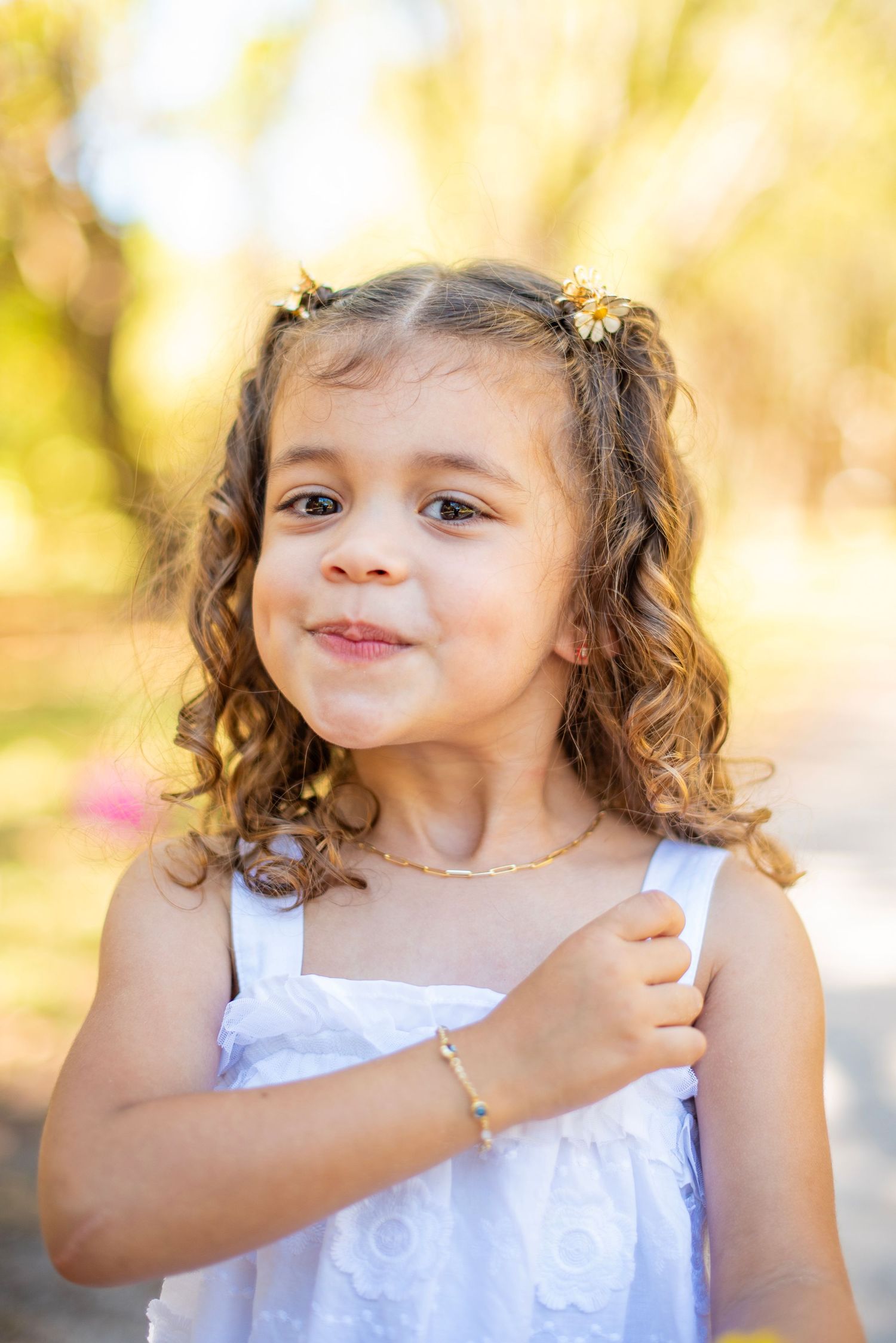 A child in a white dress poses playfully outdoors during golden hour with curly hair and a joyful expression.