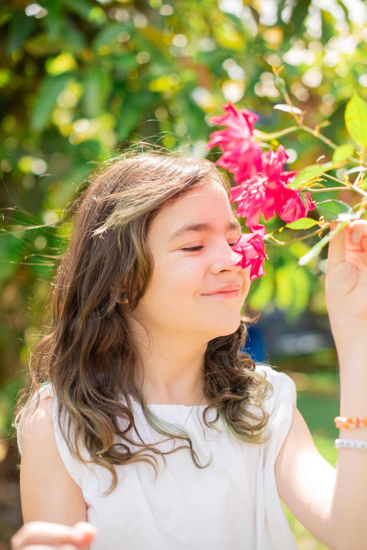 Person in white top enjoying the fragrance of pink flowers in a sunny garden setting.