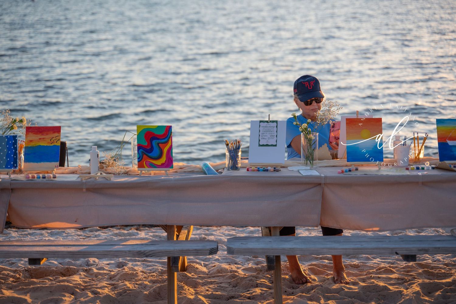 A row of colorful paintings and artwork displayed on easels along a sandy beach at sunset.