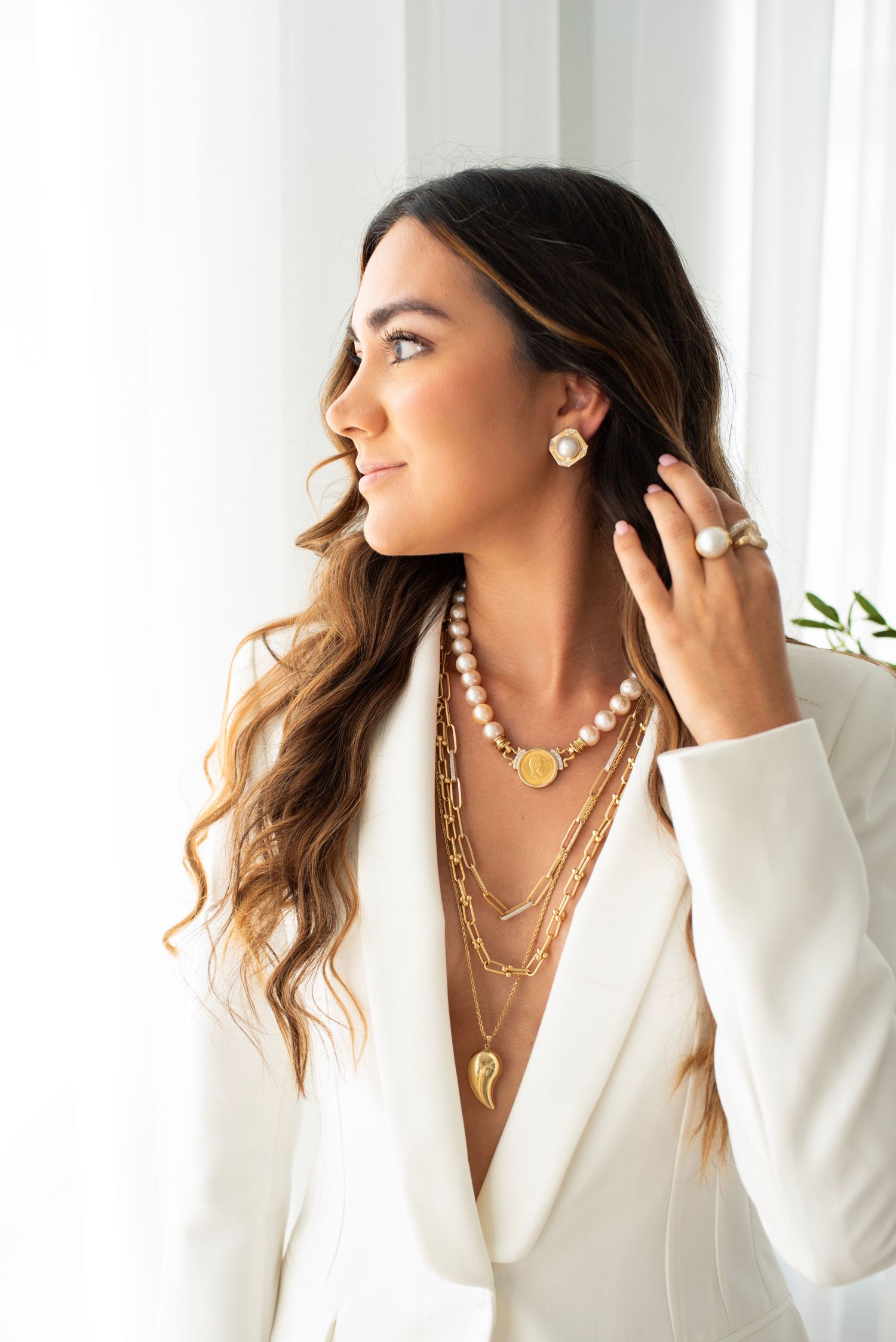 Woman in white blazer wearing layered gold necklaces and jewelry against bright background.