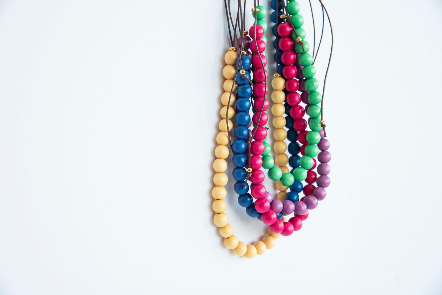 Colorful wooden bead necklaces in pink, green and gold hanging against white background.