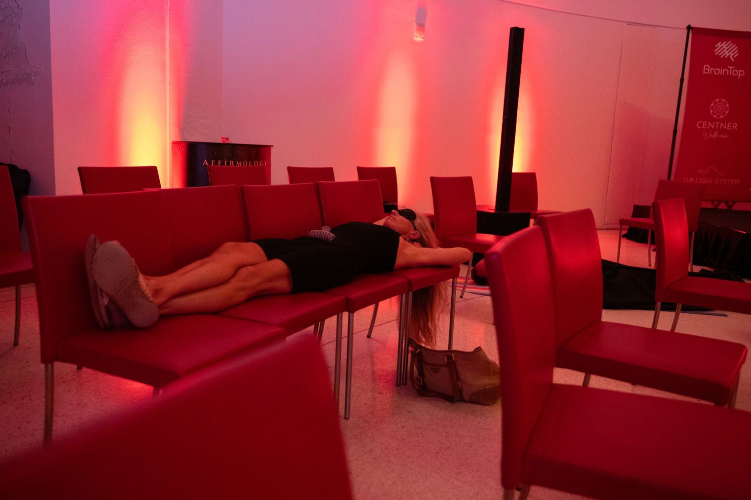 Person lying on red leather sofa in a dimly lit red room with chairs and ambient lighting.