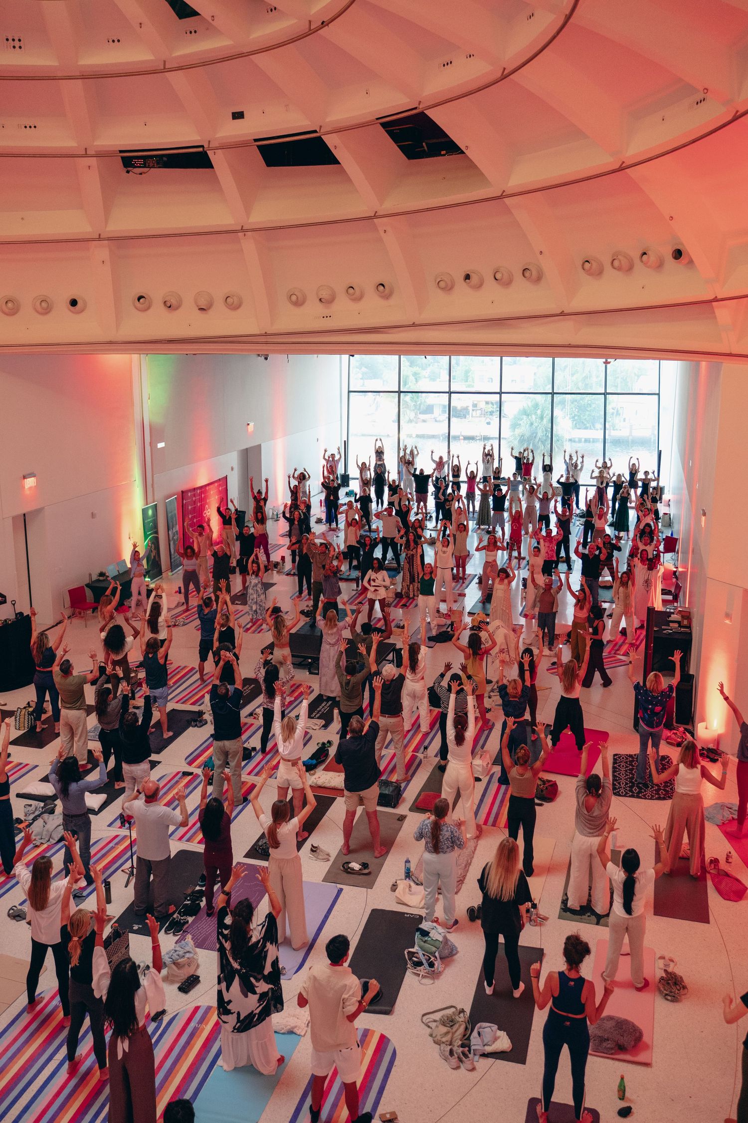 Large group yoga class in a modern white circular hall with colorful striped mats and vibrant lighting.