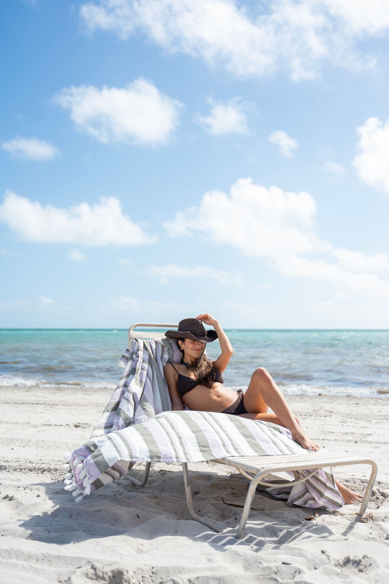 A person relaxes on a beach chair under a blue sky with turquoise ocean waters in the background.