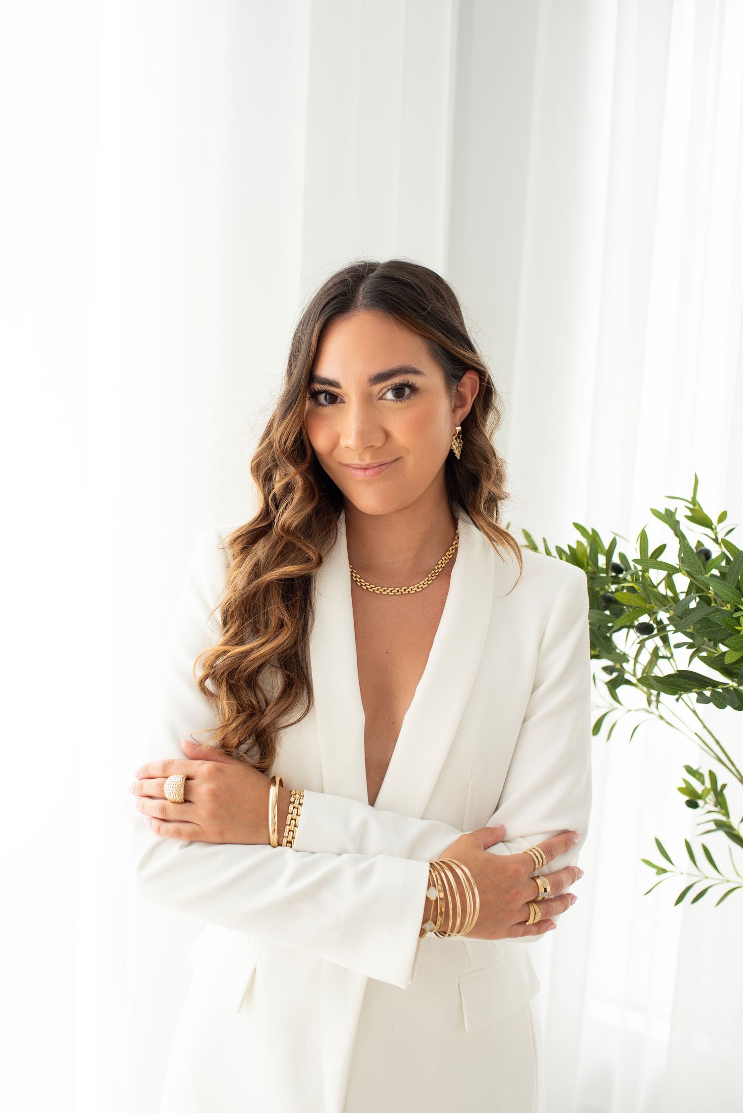 Professional portrait in a white blazer with gold jewelry and long wavy hair against a light background with greenery.