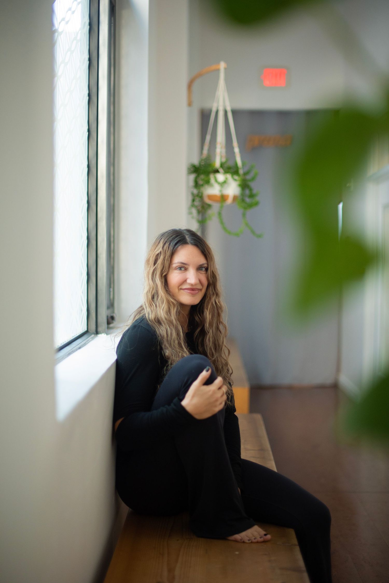 Indoor portrait in natural light with green plant leaves in foreground and window lighting creating soft ambiance.