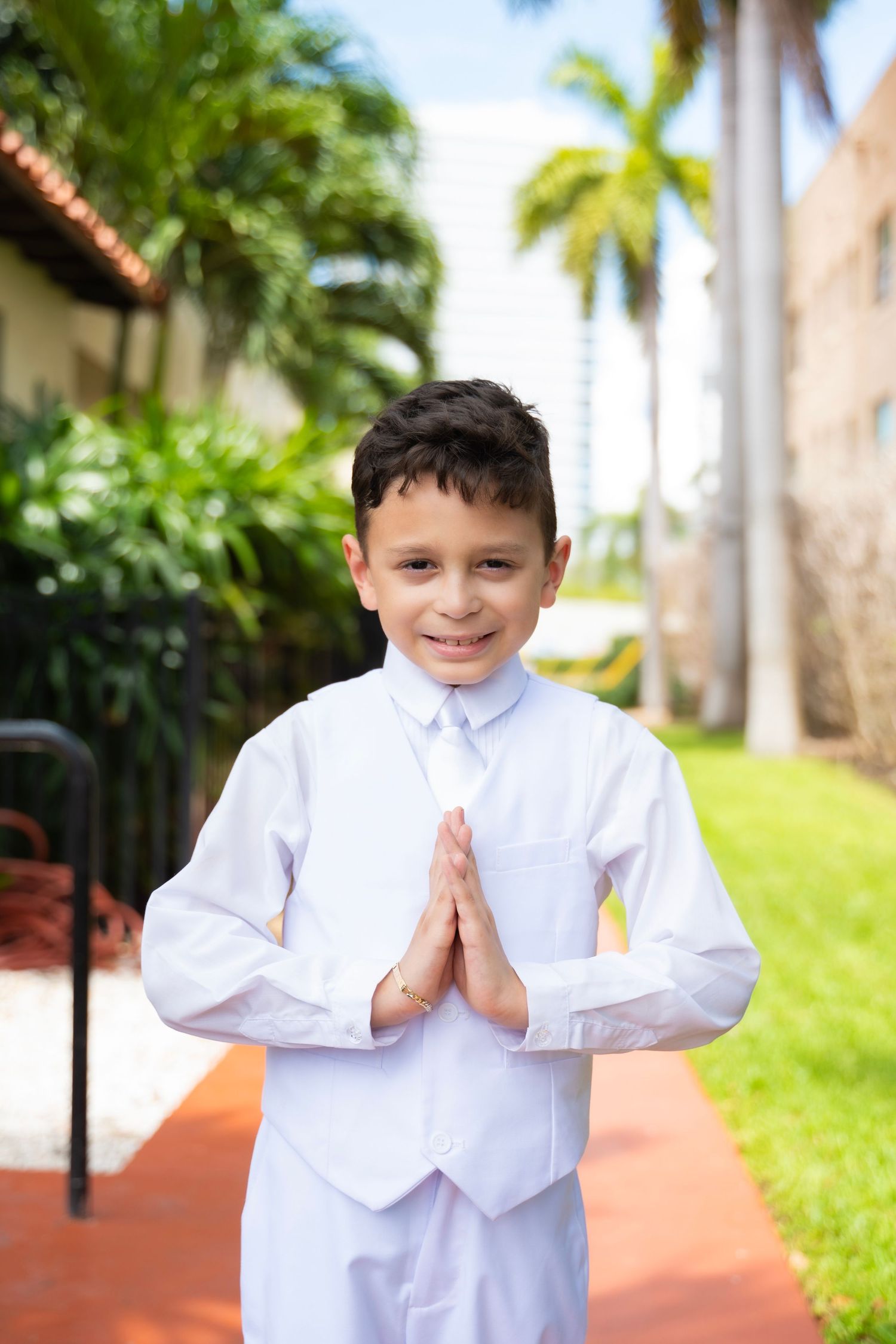 Boy in white suit with hands in prayer pose, smiling outdoors near palm trees, dressed for First Communion.