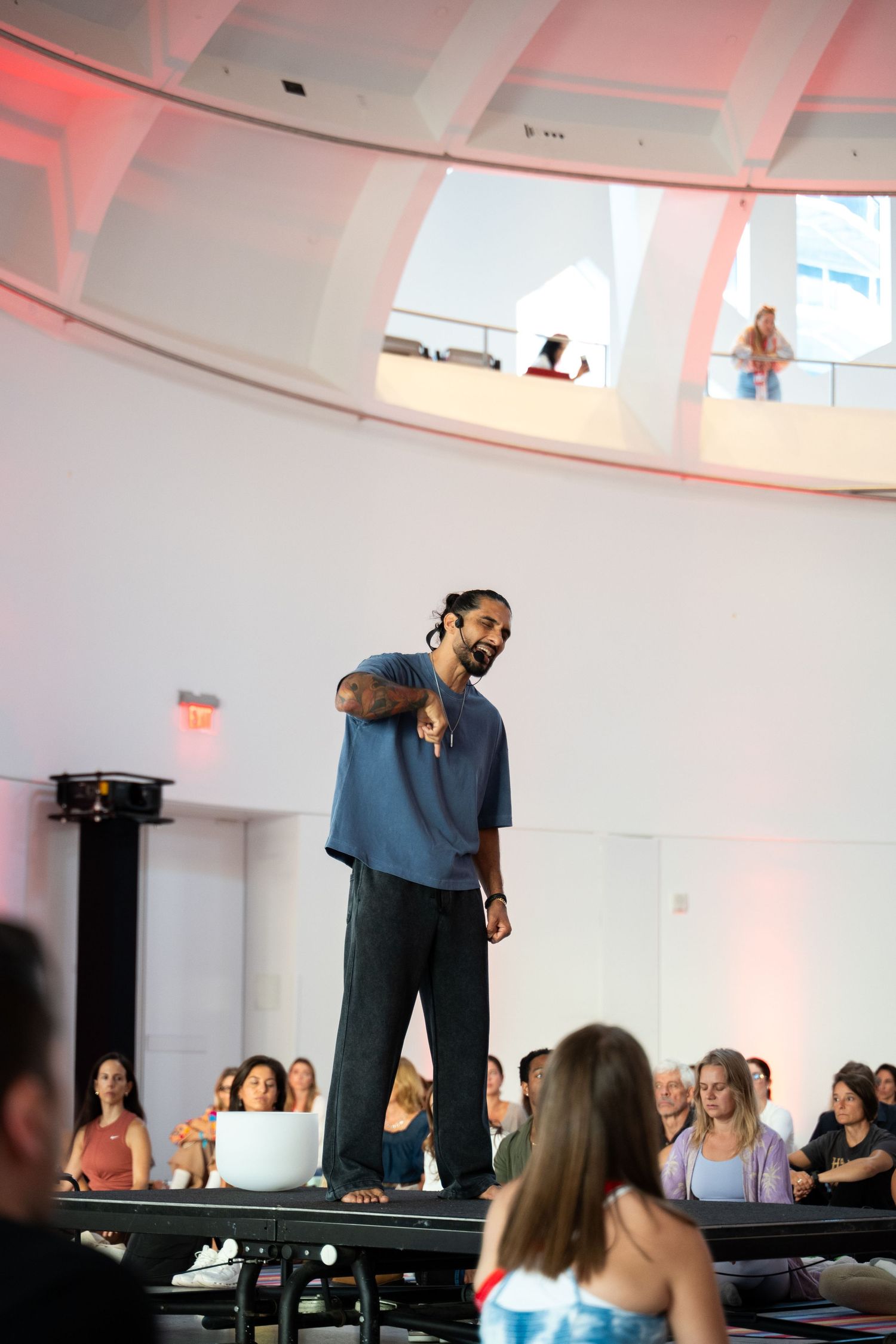 Man in blue shirt gestures to audience while speaking on stage in a modern white domed venue.