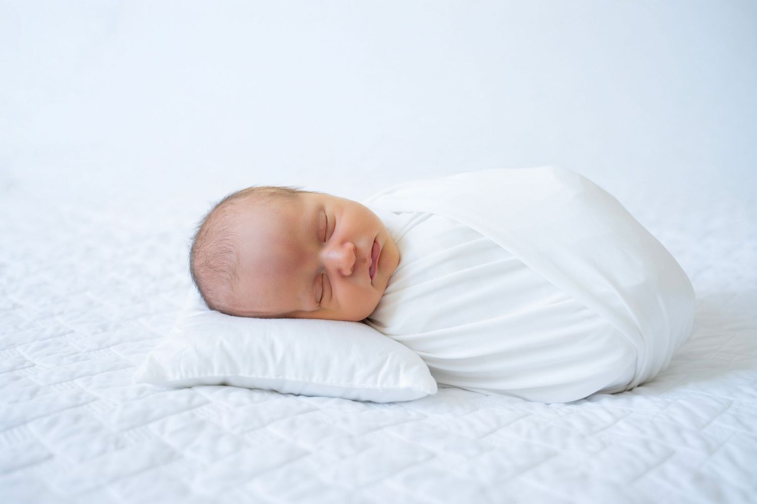 Peaceful sleeping newborn baby swaddled in white blanket rests on small pillow against white background.