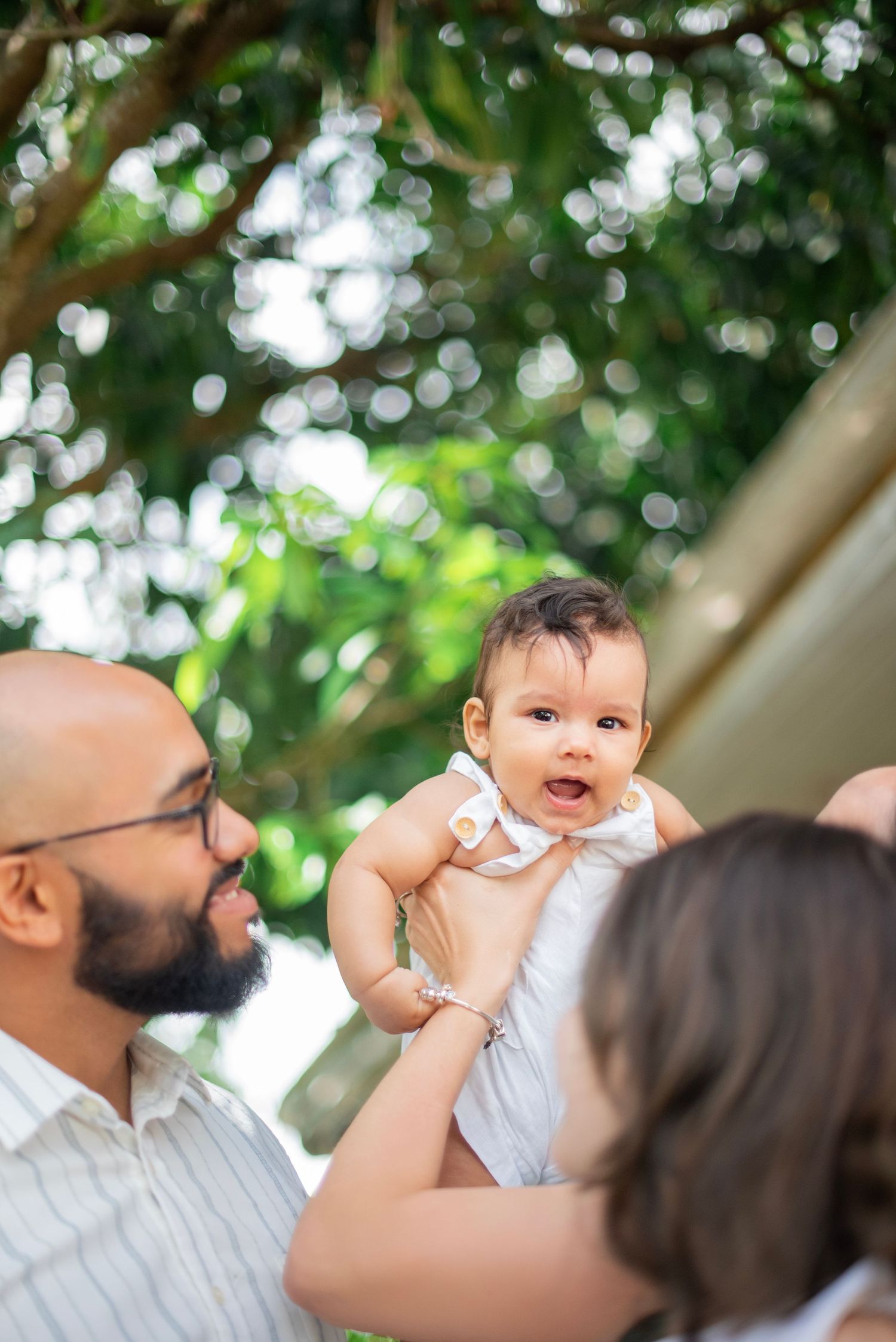 A joyful baby in white clothing smiles while being lifted up outdoors with green foliage in the background.