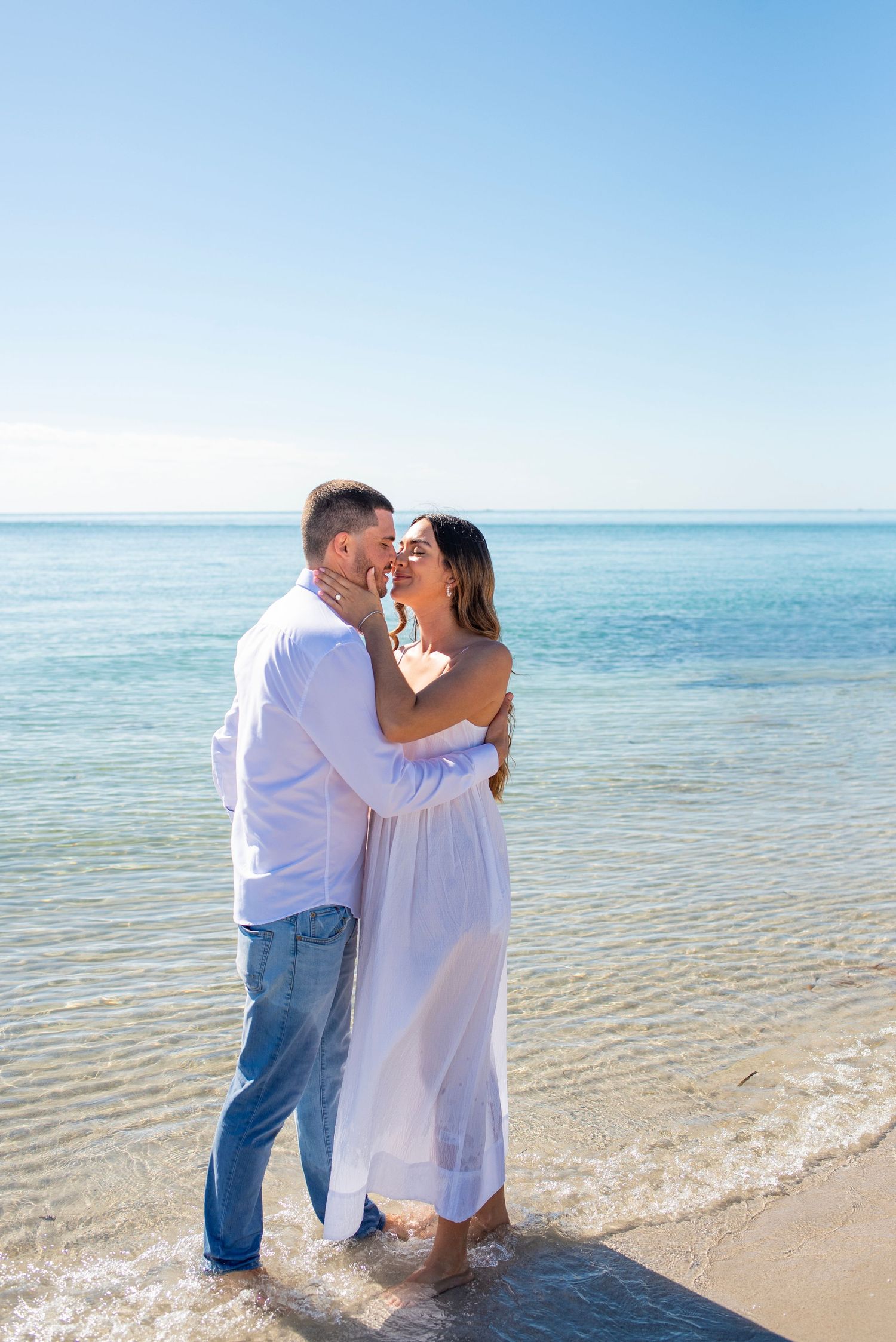 A couple in white clothing embraces romantically on a pristine beach with turquoise waters in the background.
