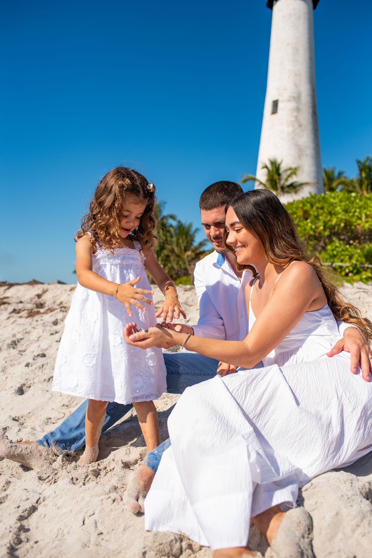 A family in white outfits poses together on a sandy beach with a lighthouse and palm trees in the background.