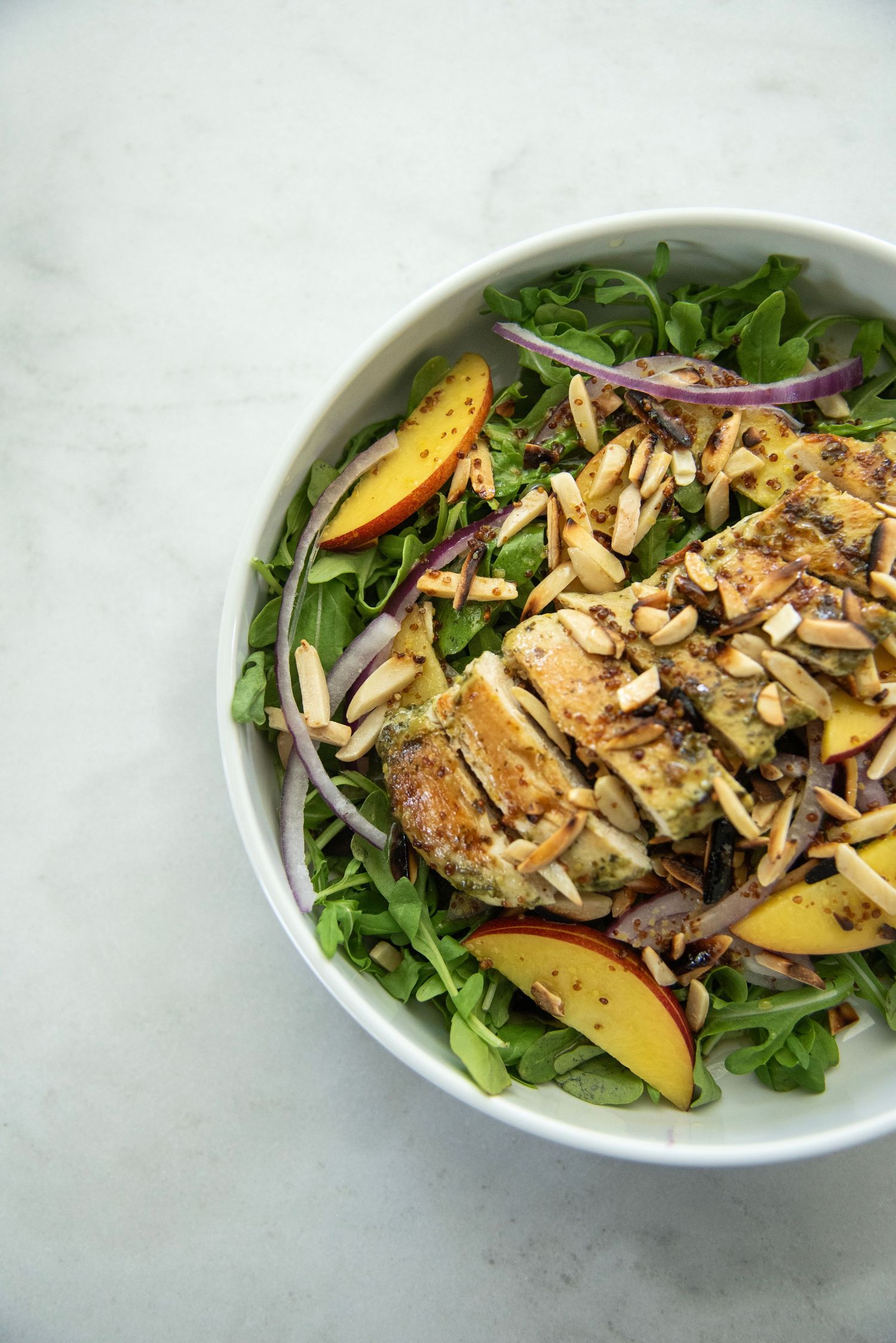Colorful salad bowl with mixed greens, sliced peaches, and toasted almonds on marble surface.