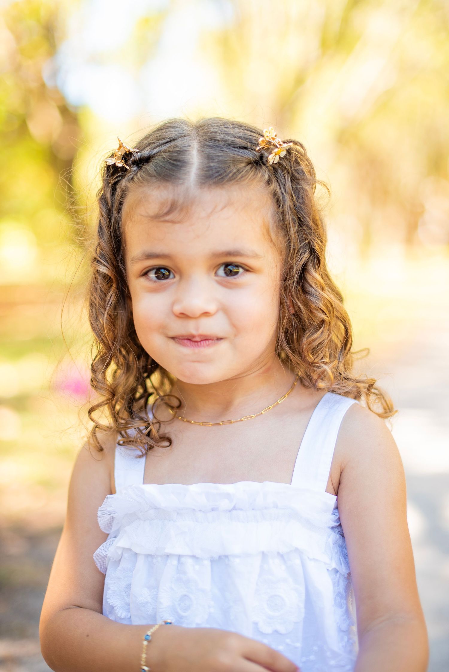 A young child in a white eyelet dress poses outdoors with soft golden sunlight creating a dreamy atmosphere.