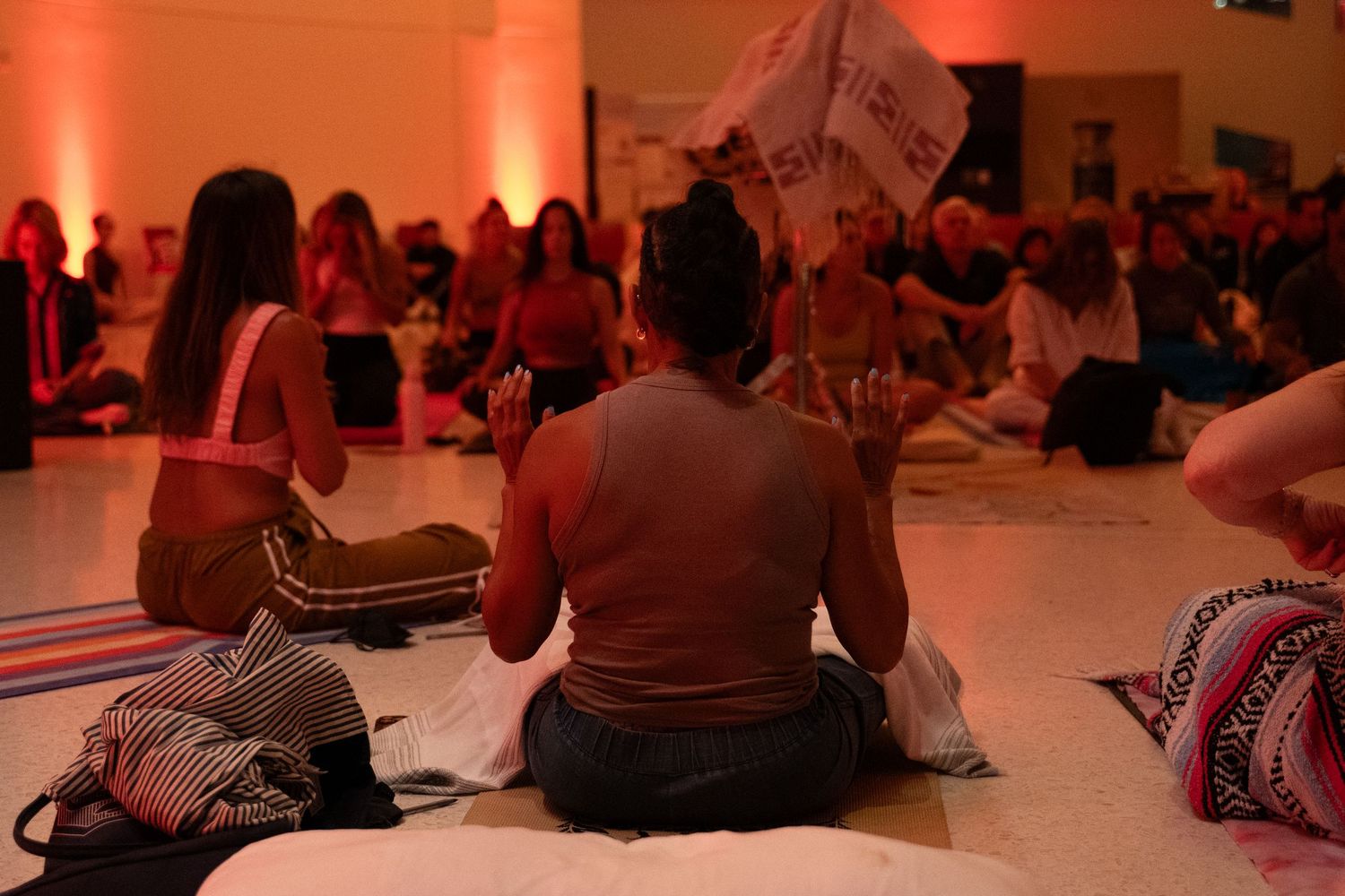 Large group of women sitting on floor at indoor wellness yoga event with warm ambient lighting.