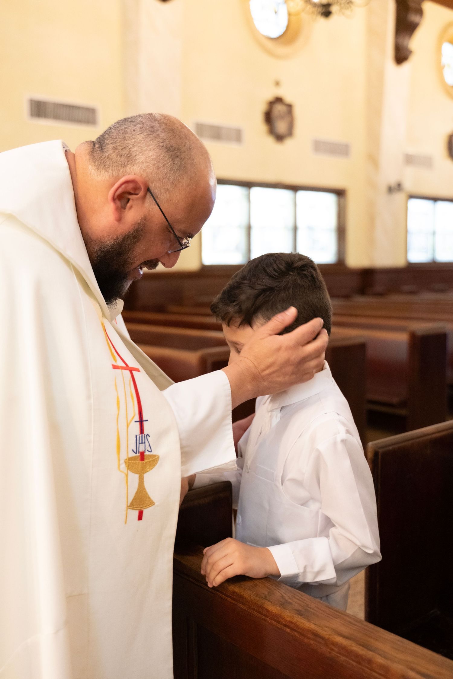 Priest in white vestments placing hand on young boy's head during a religious ceremony inside a church.