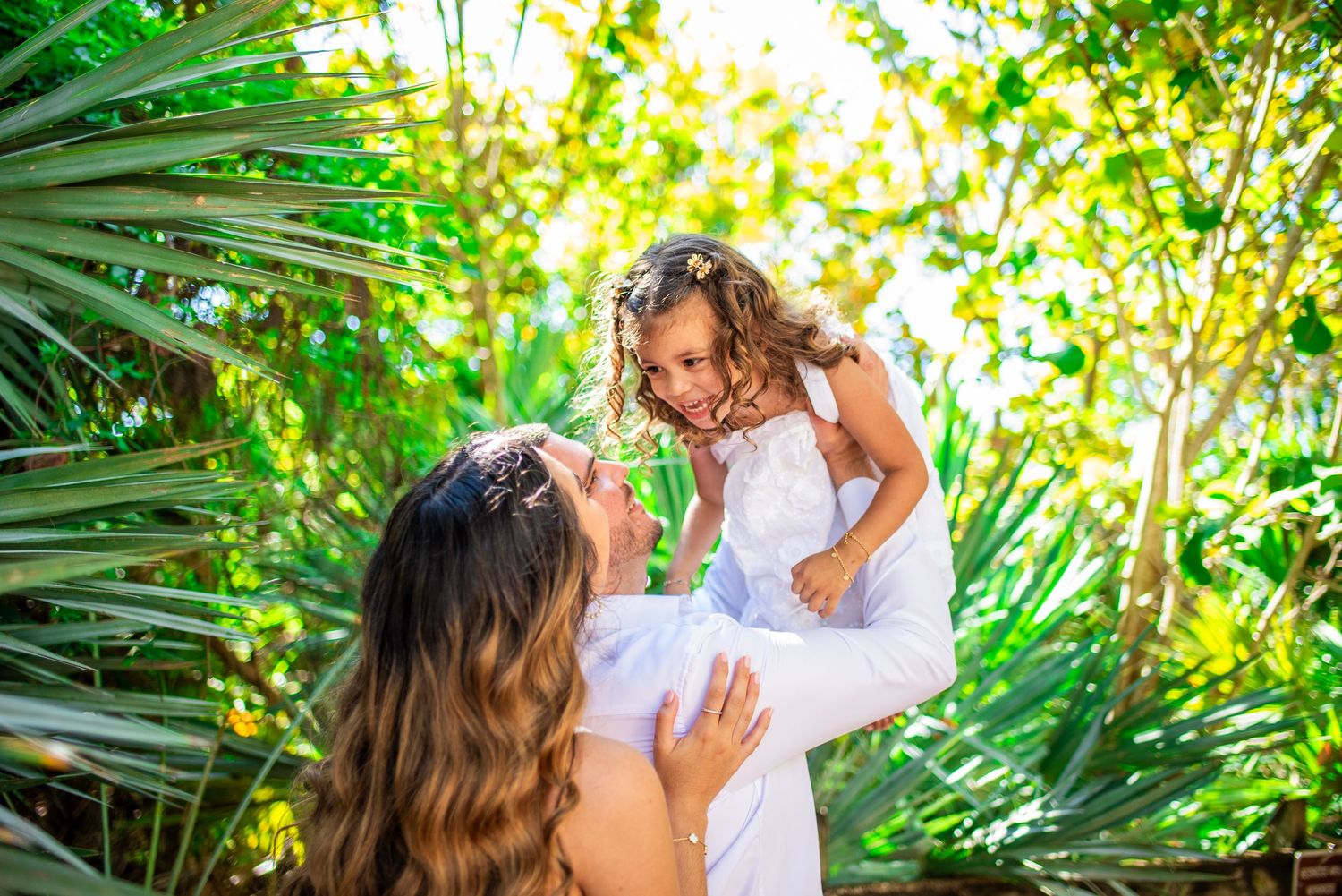 A couple embraces in a lush tropical garden setting with sunlight filtering through palm fronds and greenery.