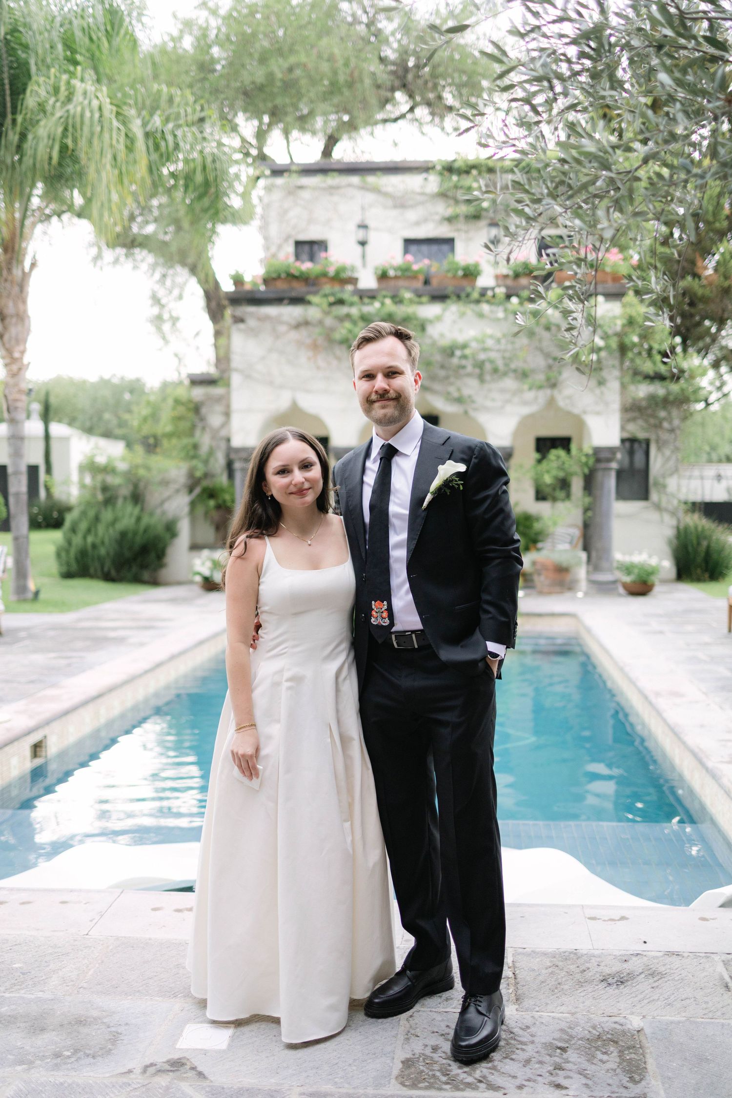 Retrato de boda en exterior en San Miguel de Allende. Pareja de novios sonrientes de pie bajo el sol, rodeados de vegetación.