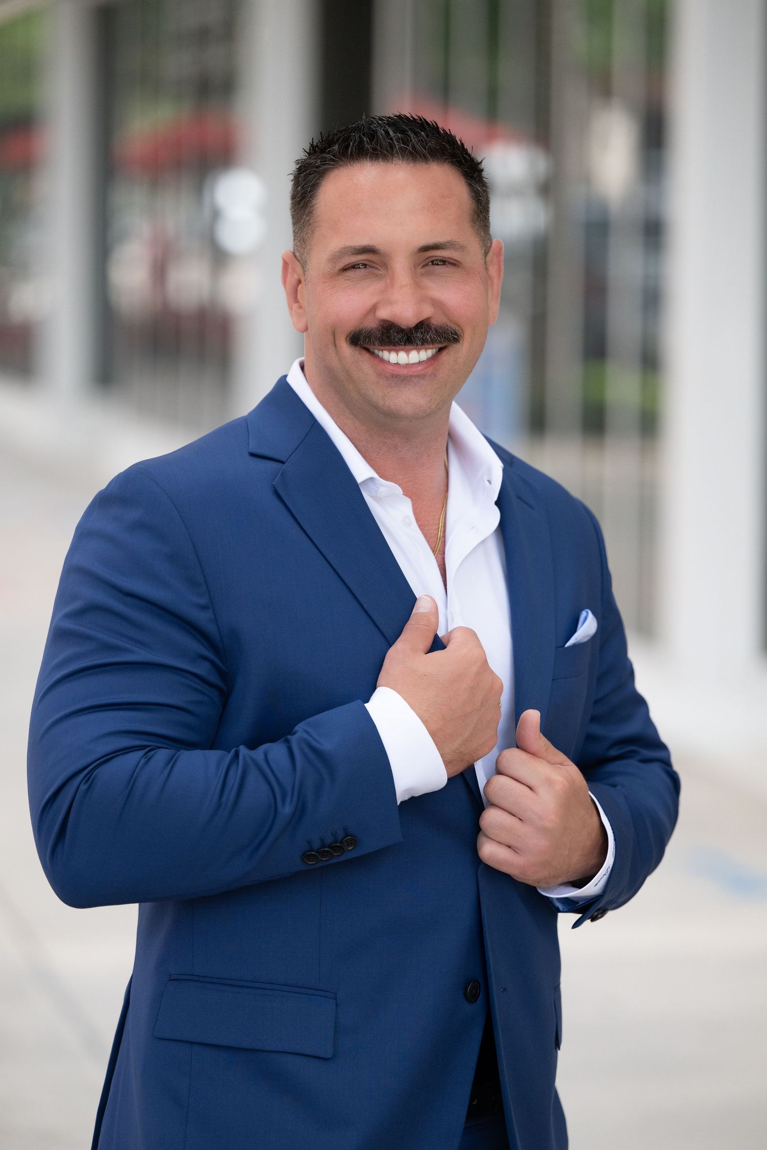 Smiling man with mustache in blue suit and white shirt posing confidently outdoors in a professional headshot.