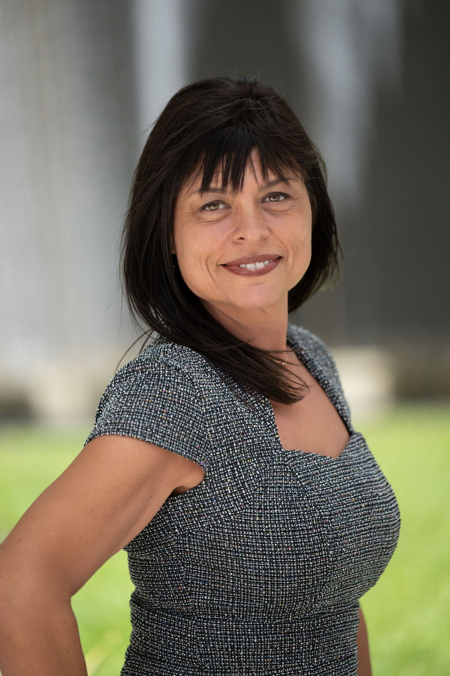 Professional woman with dark hair smiling outdoors in a gray dress, posing confidently for a business portrait.