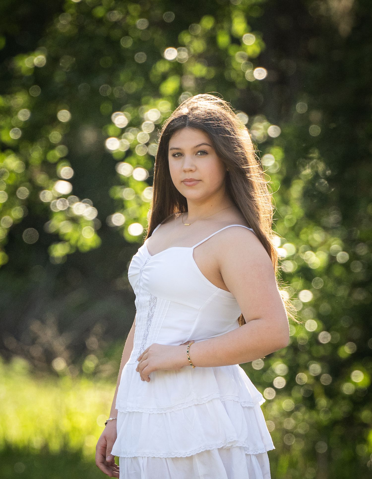 A person in a flowing white sundress poses gracefully in a sun-dappled outdoor setting with bokeh lighting effects.