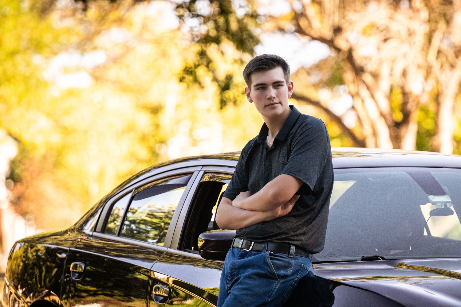A person in a black polo shirt stands beside a dark luxury car against a golden sunset backdrop.