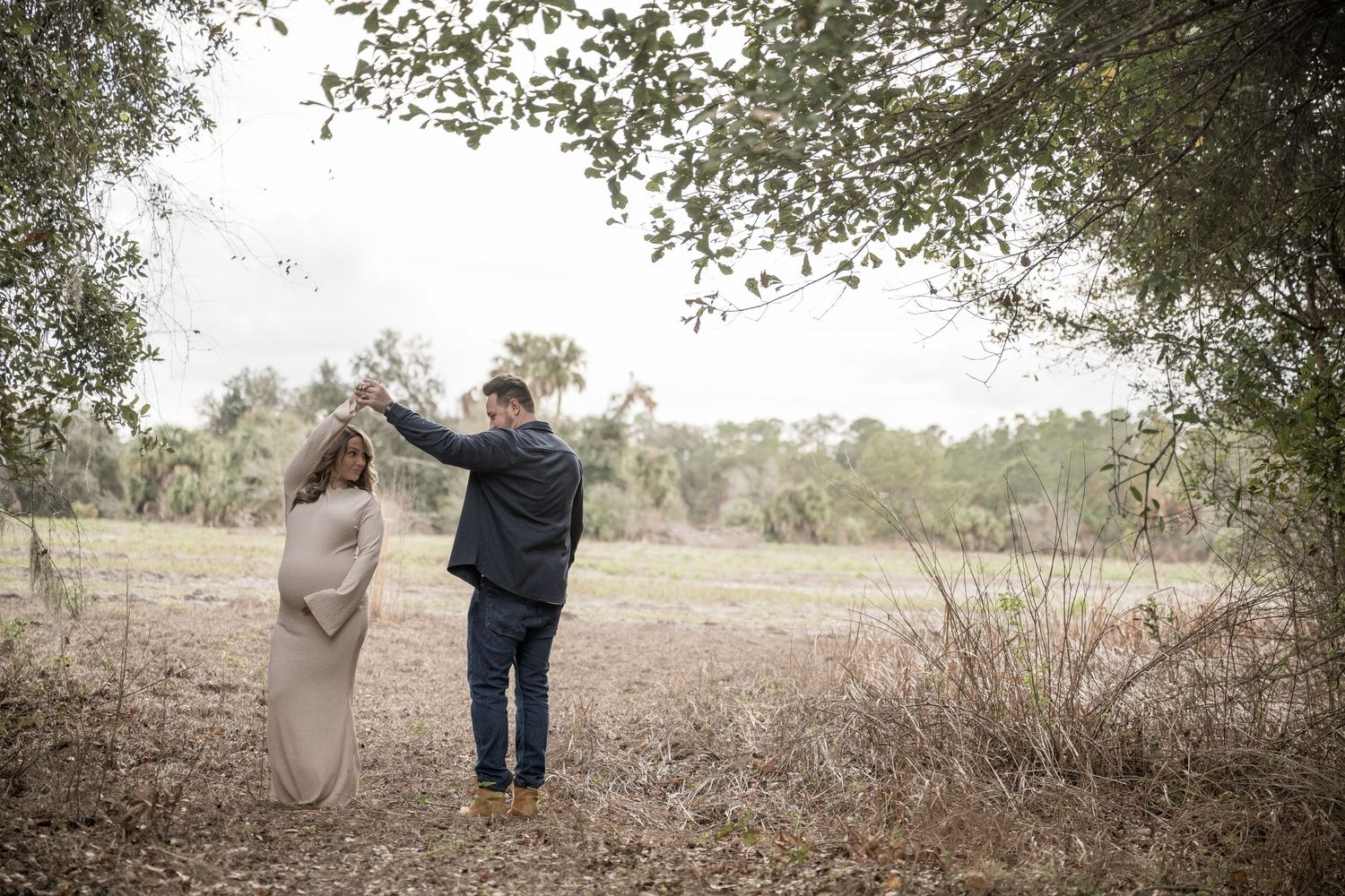A couple in formal attire dances together in a rustic outdoor setting surrounded by trees and natural landscape.