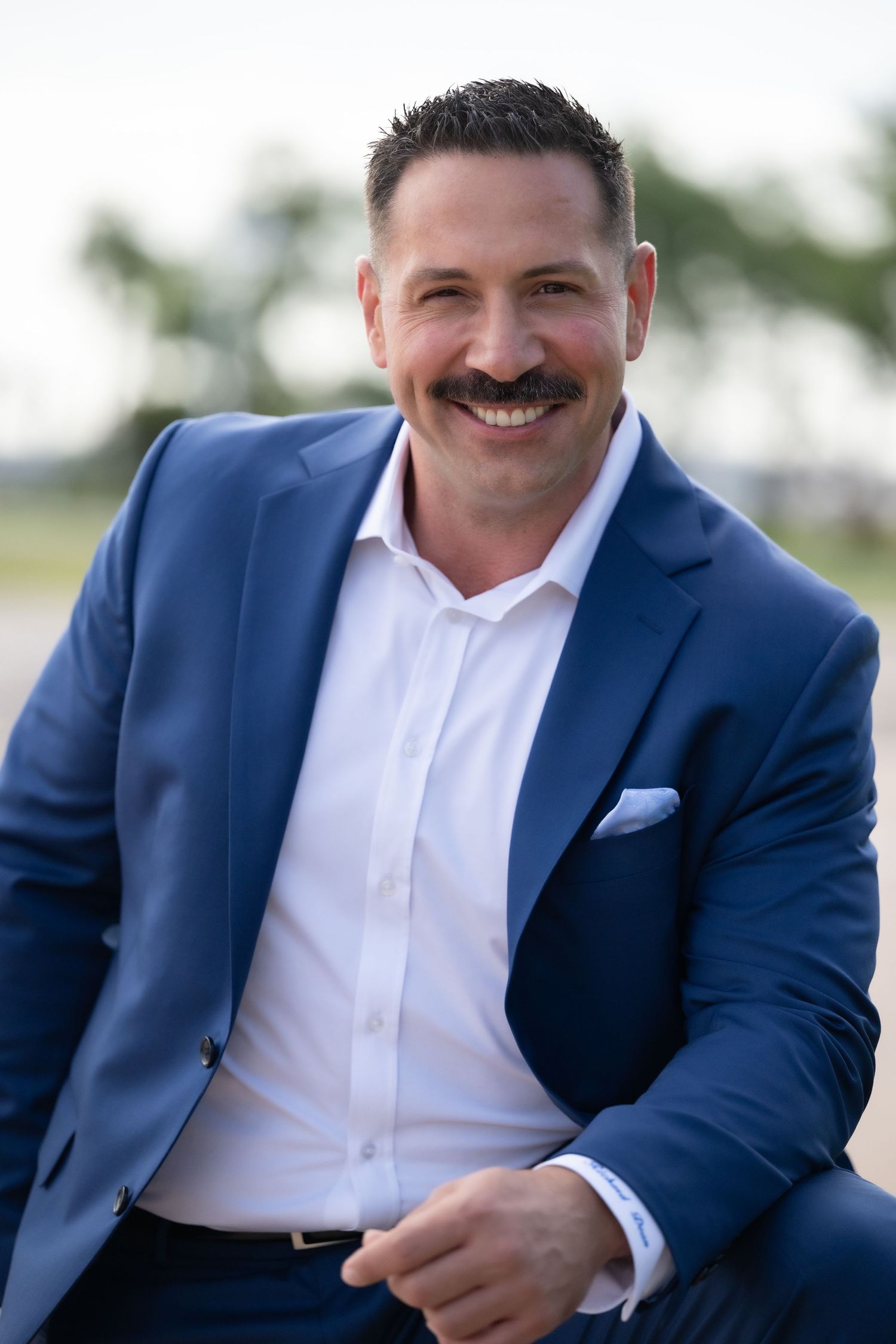 Smiling man in blue blazer and white shirt with mustache poses outdoors in professional headshot photo.