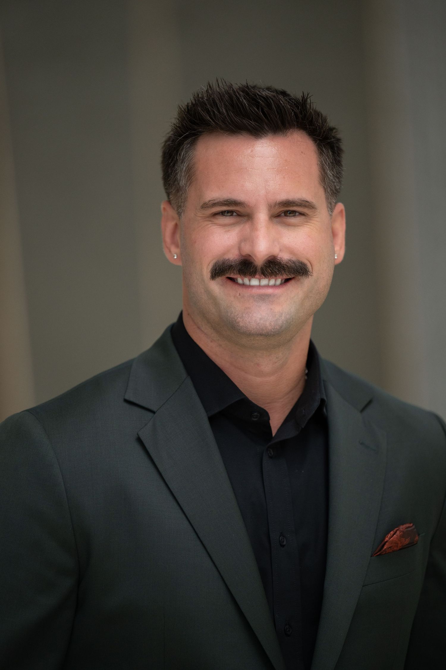 Professional man with mustache smiling in dark green blazer and black shirt with red pocket square.