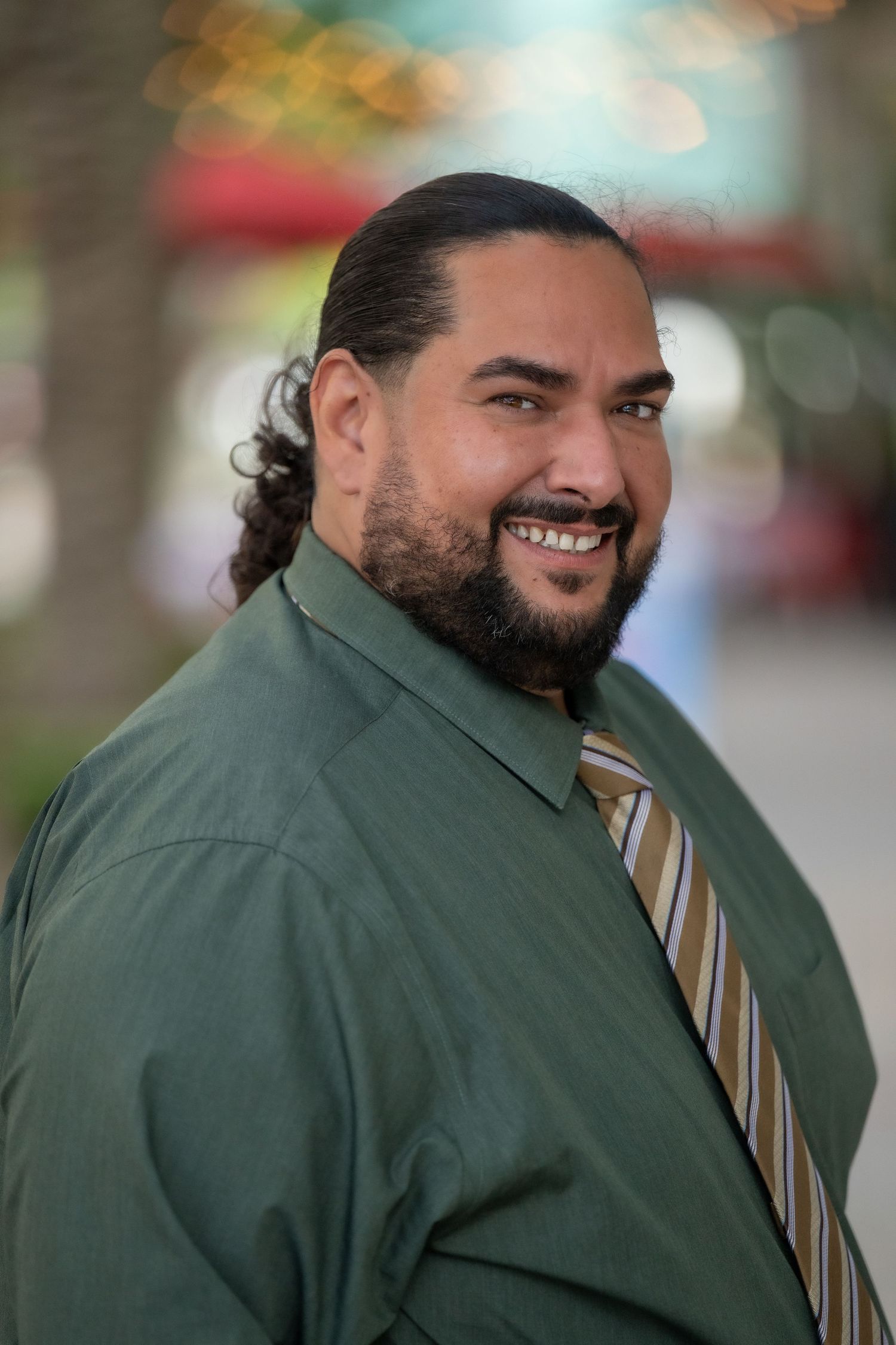 Smiling bearded man with long hair in a green shirt and striped tie poses outdoors in a blurred urban setting.
