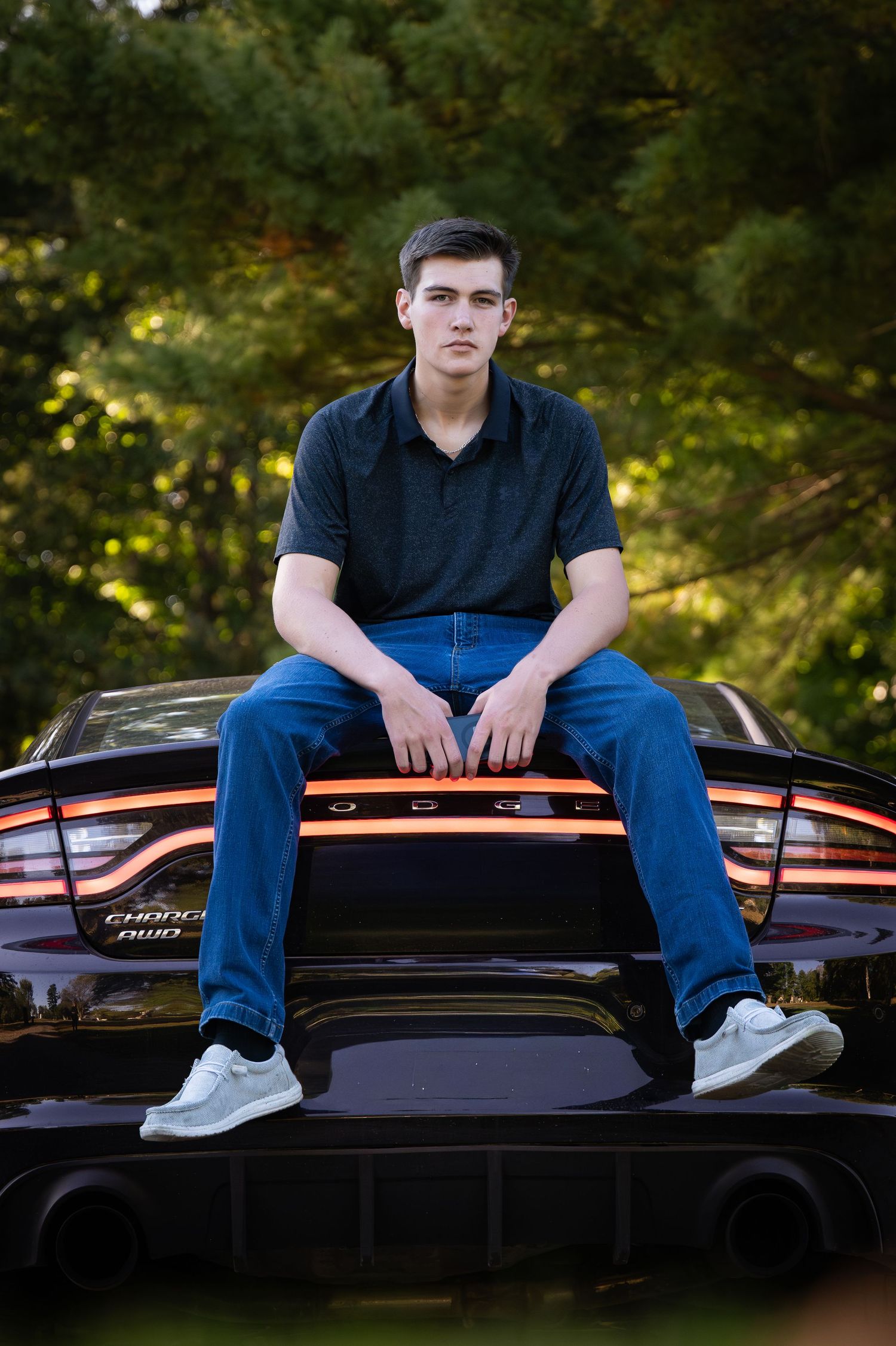 Person in navy blue shirt and jeans sits casually on top of a black car in an outdoor setting with trees in background.