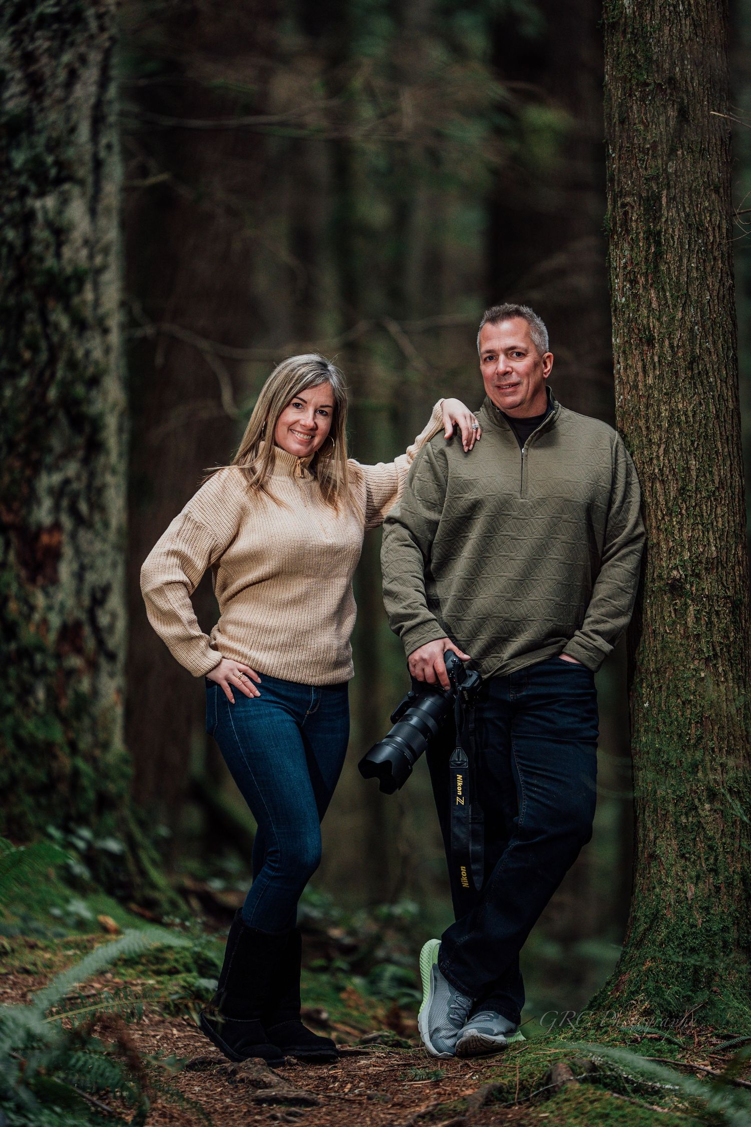 A couple in casual sweaters poses playfully against trees in a moody forest setting during an outdoor photoshoot.