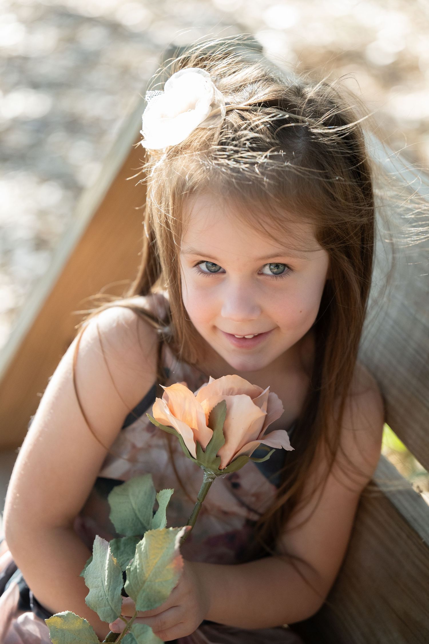 Young girl with long brown hair and blue eyes holds a peach rose, wearing a floral dress with a white flower hair clip.