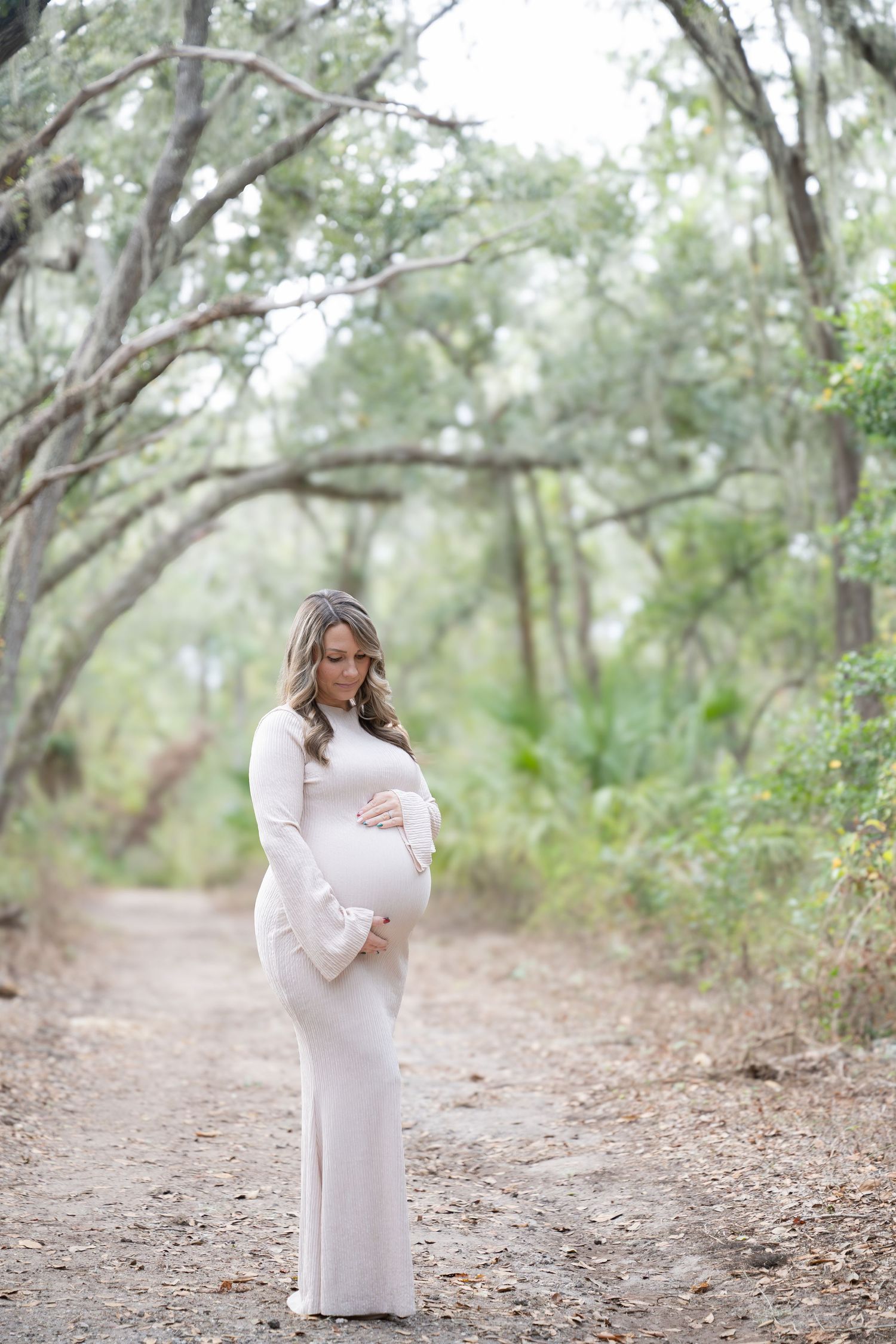 A person in a white maternity dress poses gracefully on a wooded path surrounded by eucalyptus trees.