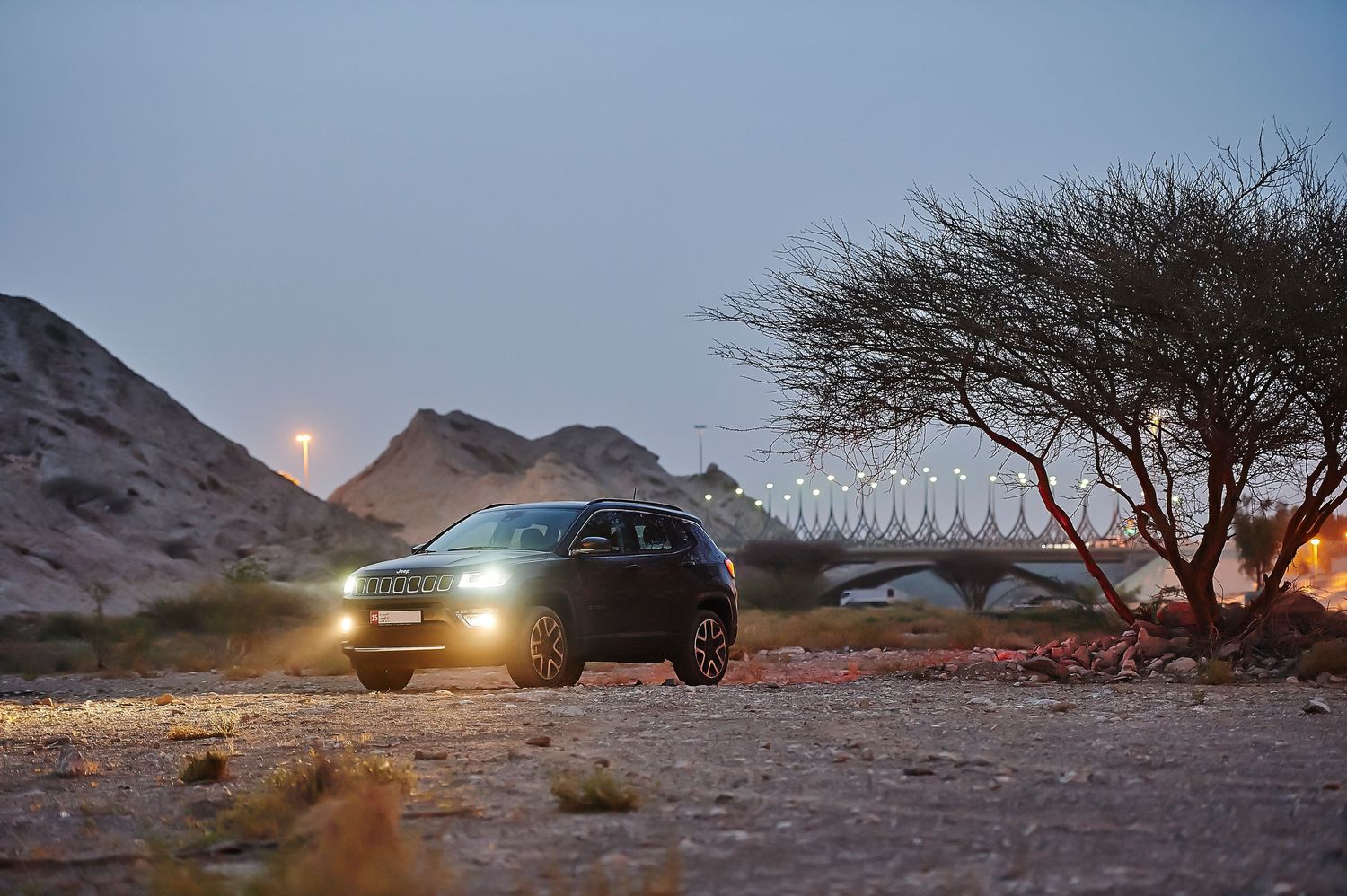 Jeep Compass SUV with headlights on at dusk in desert landscape with tree silhouette.