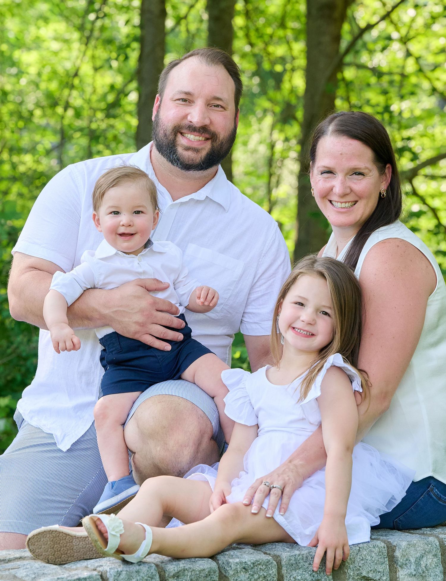 A family in white clothing sits together on rocks surrounded by lush green trees during an outdoor photoshoot.