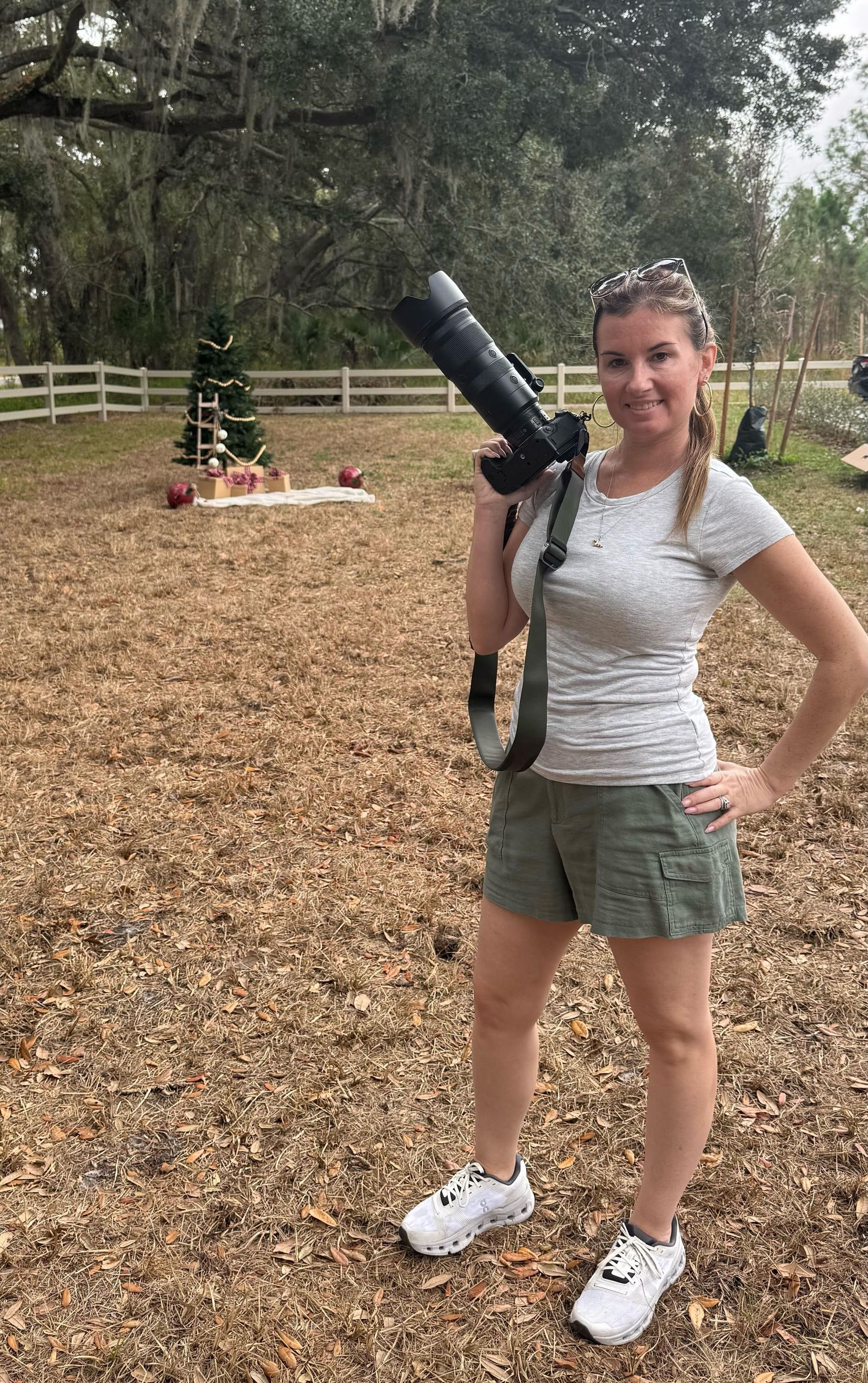 A person in a white t-shirt and green shorts stands outdoors holding a camera in a dirt field area.
