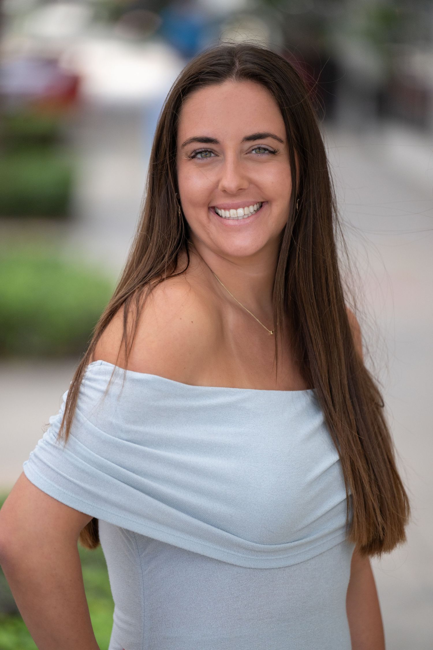 Young woman with long brown hair smiling outdoors in a light blue off-shoulder top.