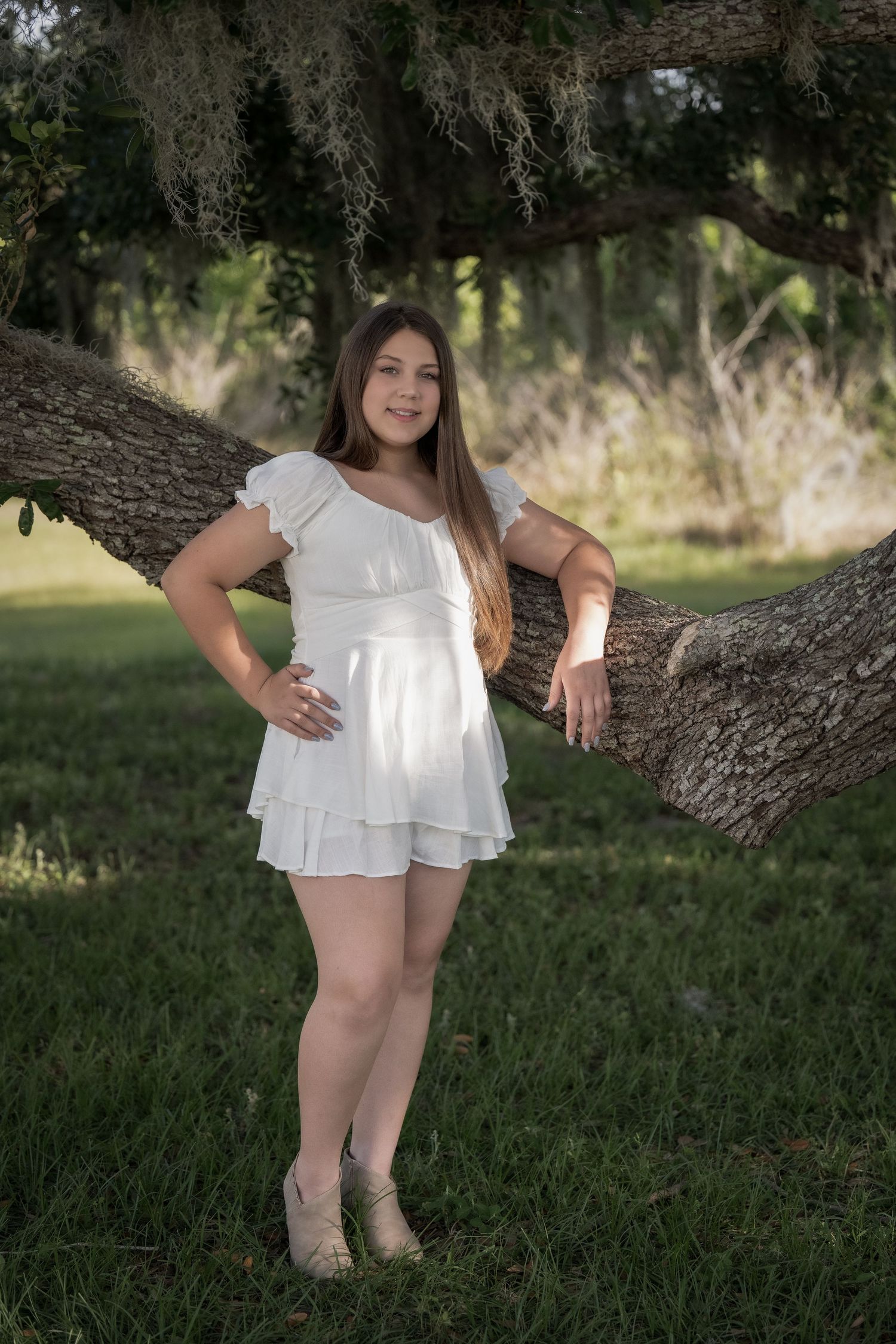 A young child in a white dress poses playfully near a large tree trunk in a grassy outdoor setting.