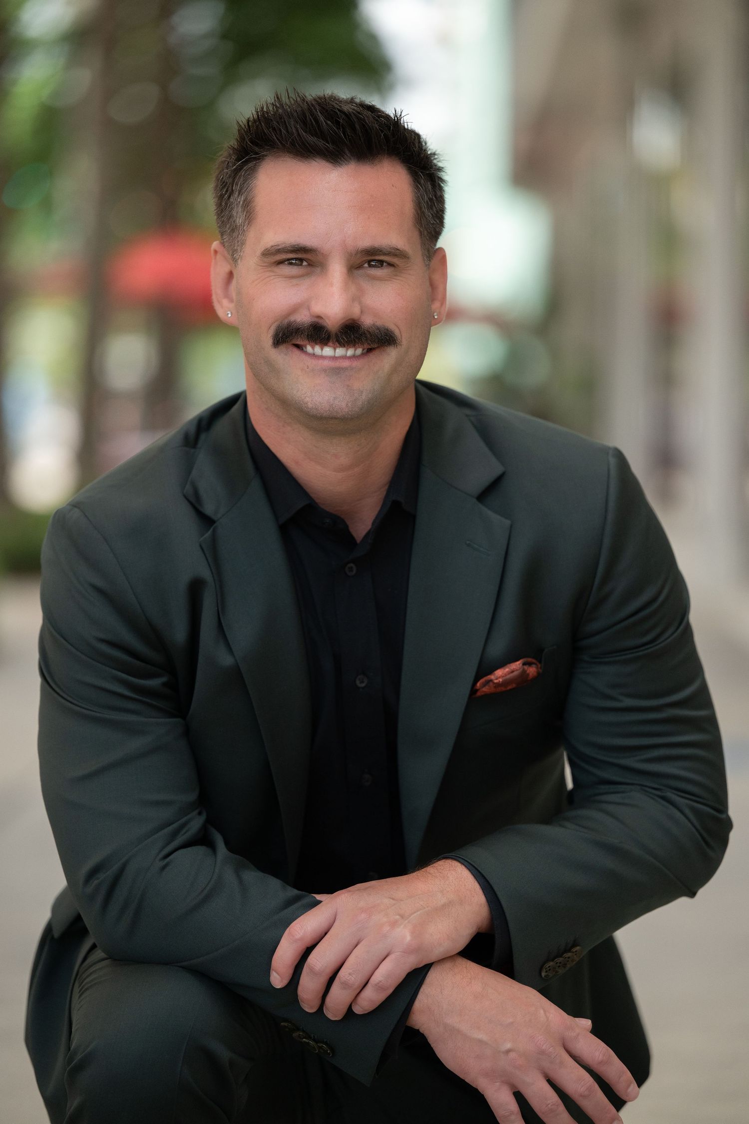 Professional man with mustache smiling in dark green suit with red pocket square, seated outdoors in urban setting.