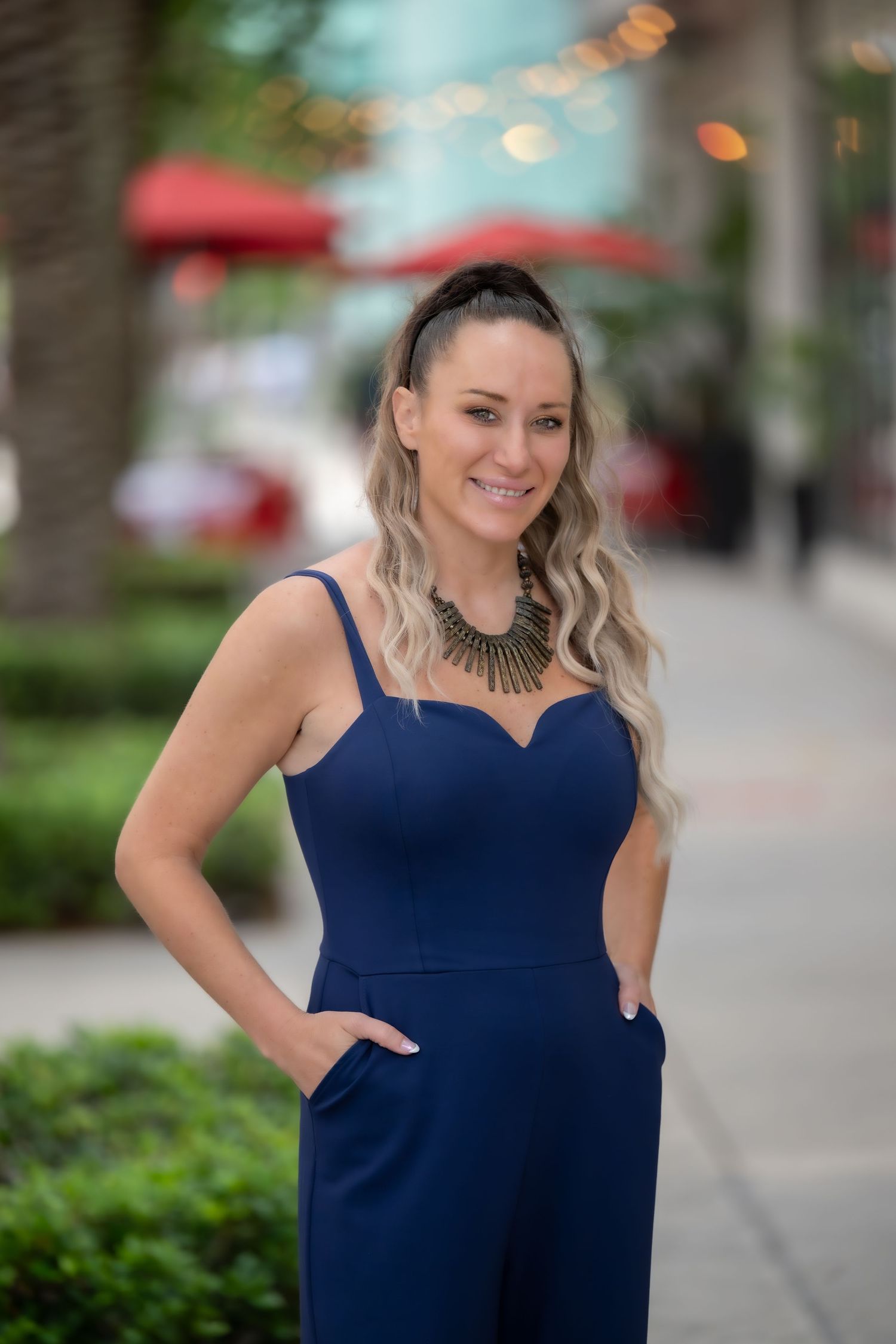 Woman in navy blue jumpsuit with statement necklace smiling outdoors on urban street with blurred background.
