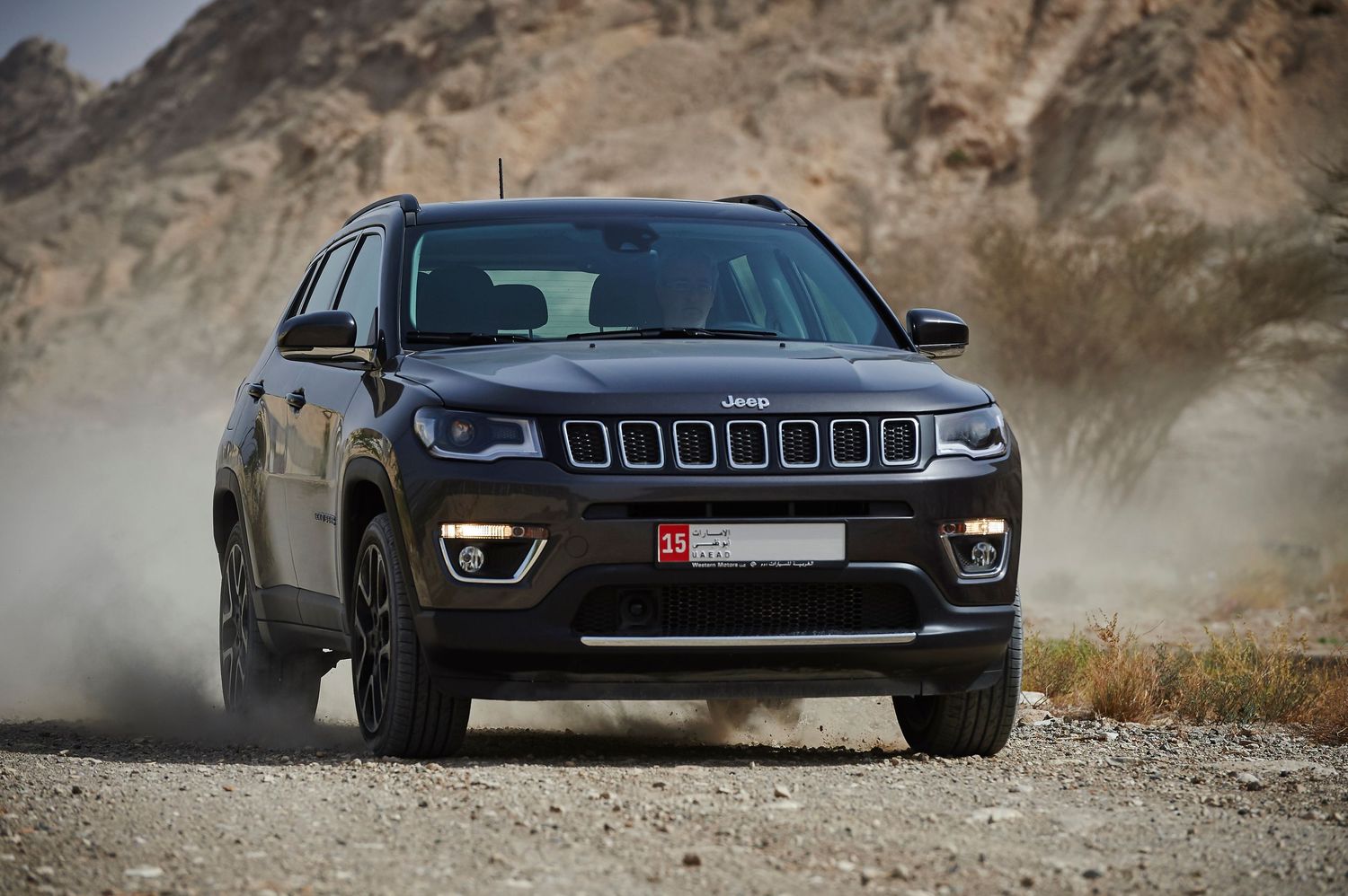 Jeep Compass SUV driving on dusty mountain road with dramatic rocky backdrop.