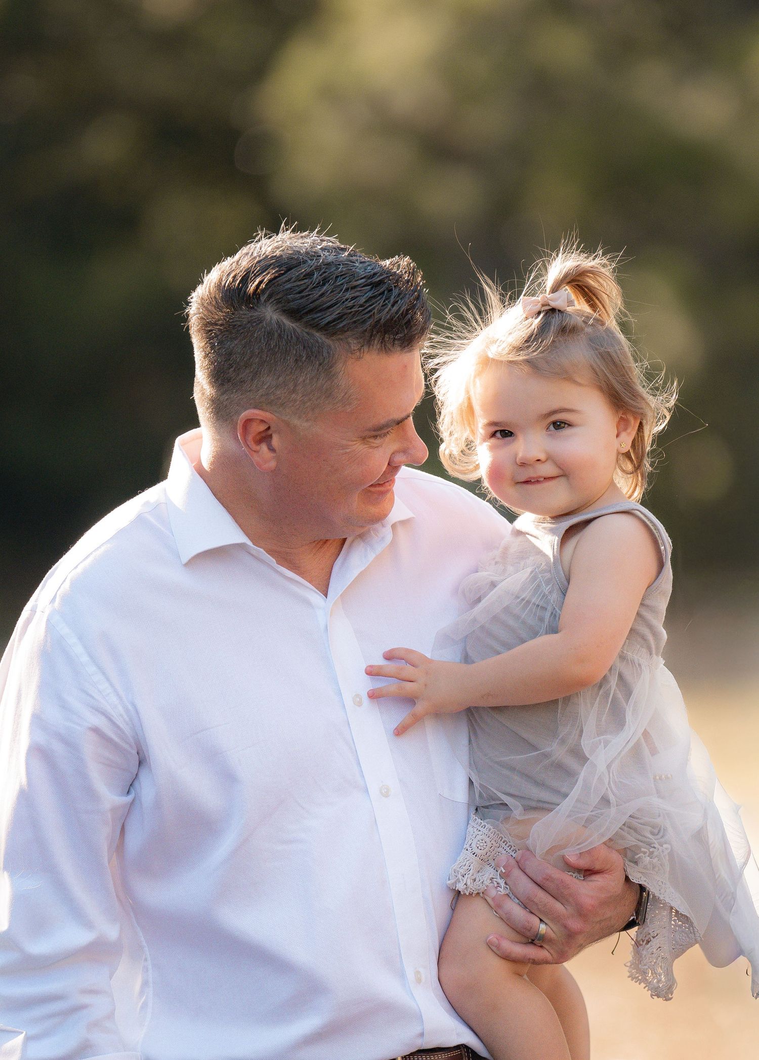 A parent in a white shirt shares a tender moment with a toddler wearing a light-colored dress in outdoor sunlit setting.