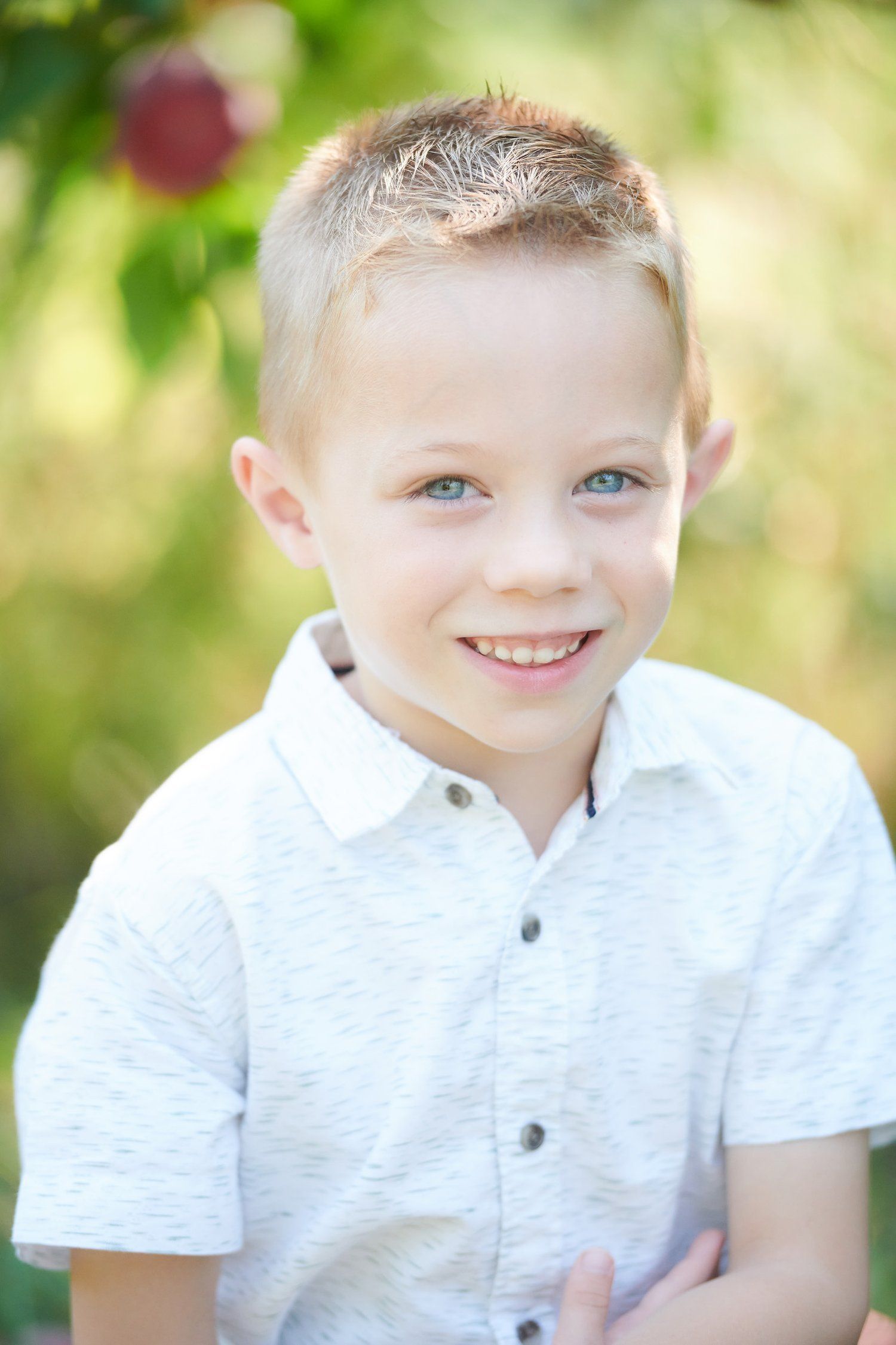 Child wearing a white collared shirt smiles while posing outdoors against a soft green natural background.